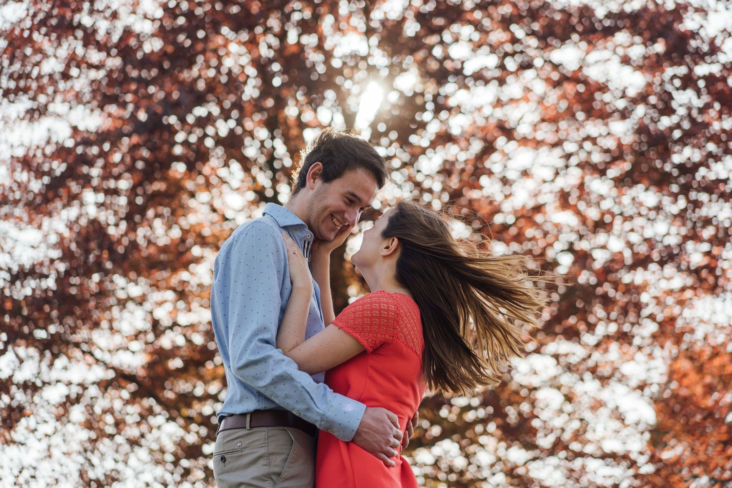 A couple embraces outdoors in autumn with red and orange fall foliage in the background.