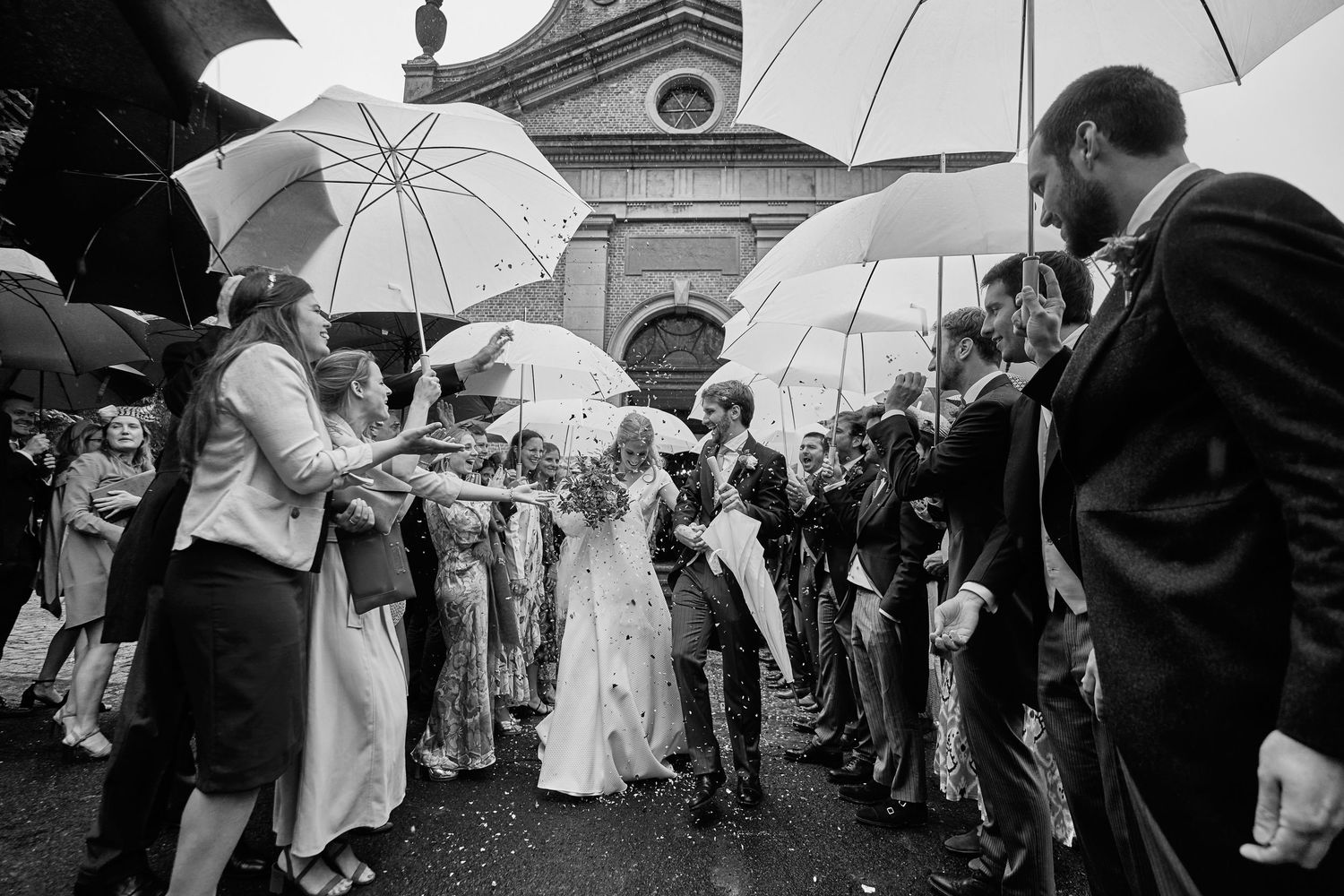 Wedding guests hold white umbrellas while gathered outside a historic building during a rainy celebration.