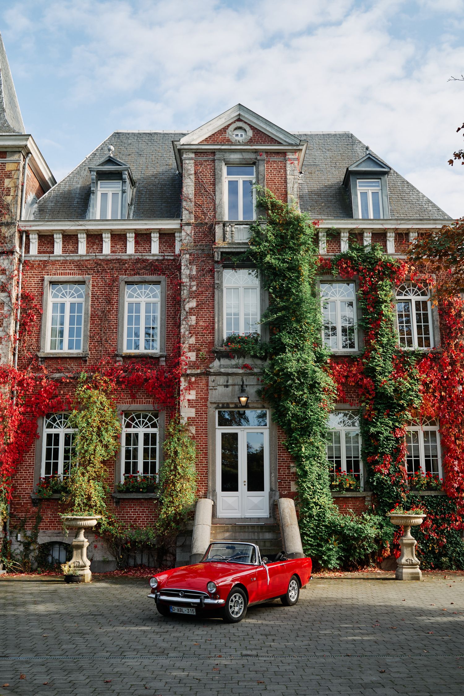 A vintage red convertible sports car parked in front of a grand brick mansion with climbing vines and fall foliage.