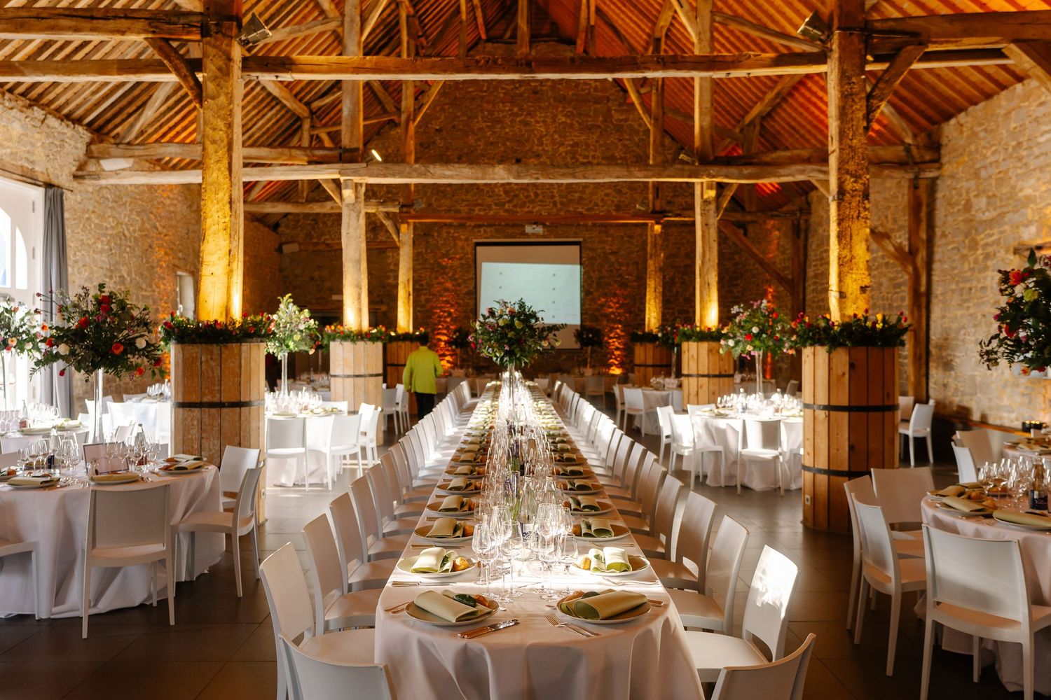 Rustic barn wedding venue with exposed wooden beams, white chairs, and elegant table settings illuminated by warm lighting.