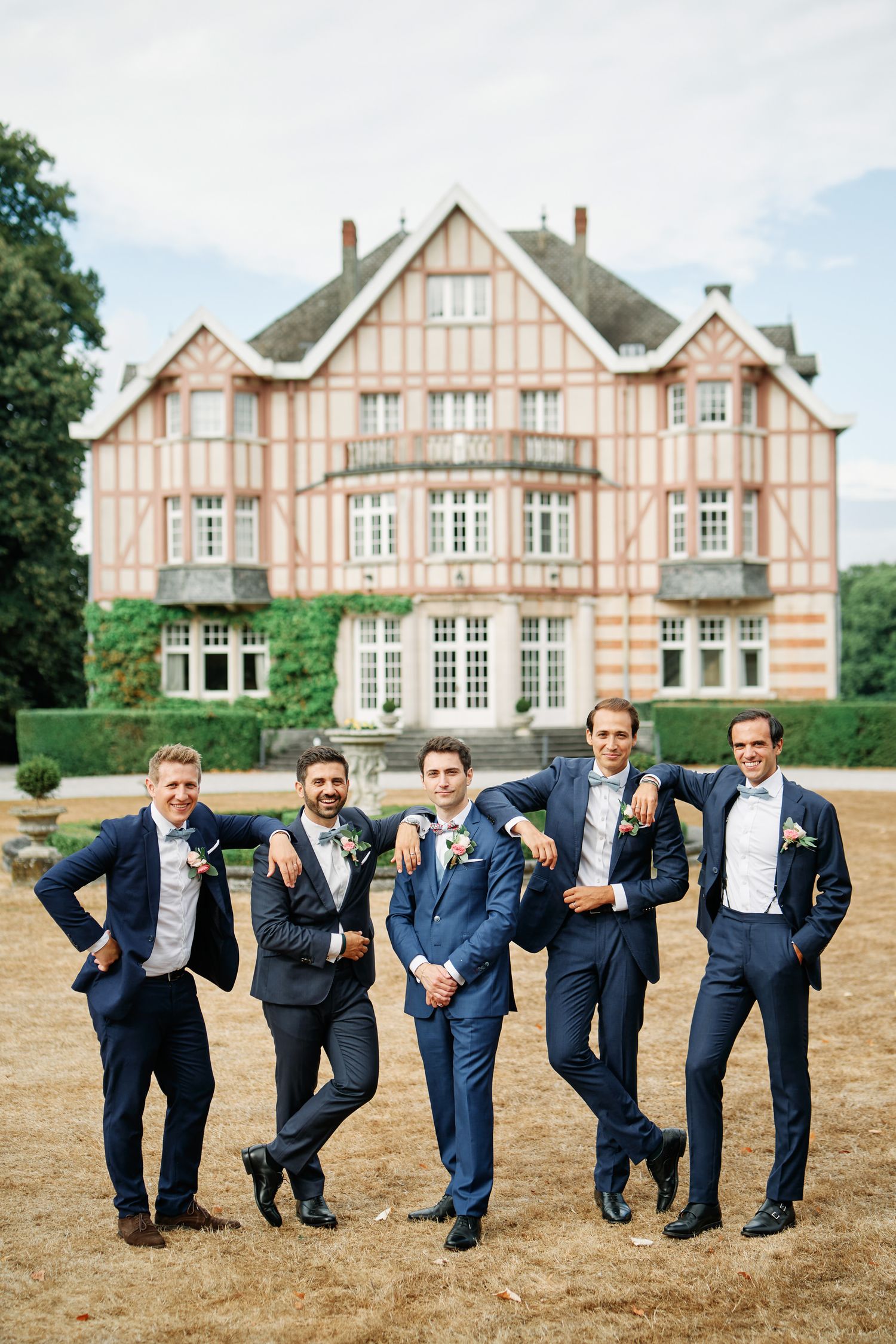 Groomsmen in navy suits pose playfully in a line on lawn with Tudor-style mansion in background.
