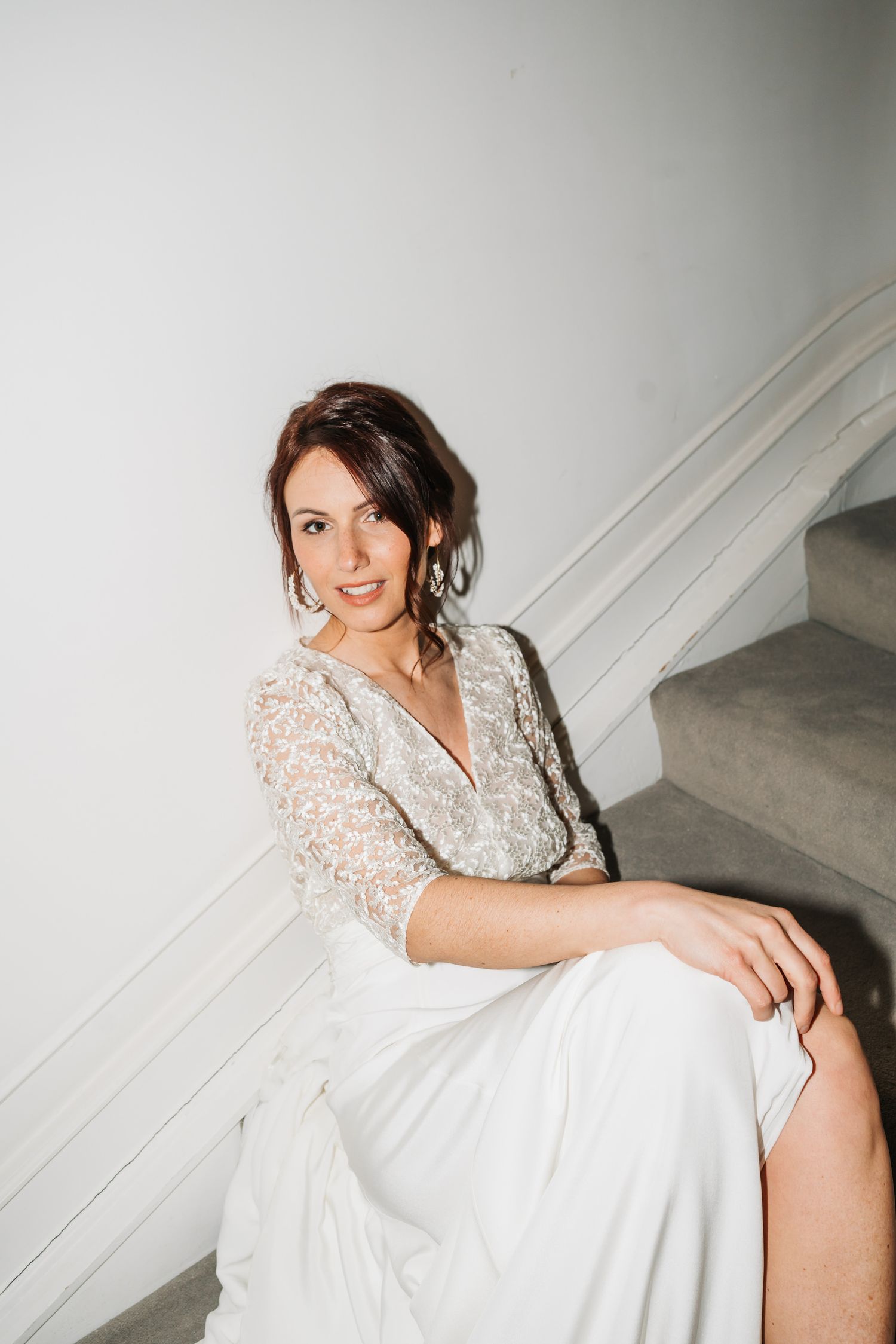A person in a white lace dress sits elegantly on white stairs in a romantic bridal portrait.