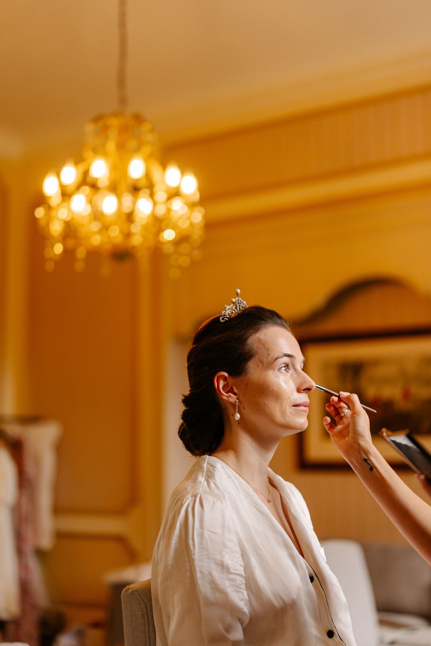A makeup artist applies cosmetics to someone wearing a white robe in a warmly lit room with chandeliers.