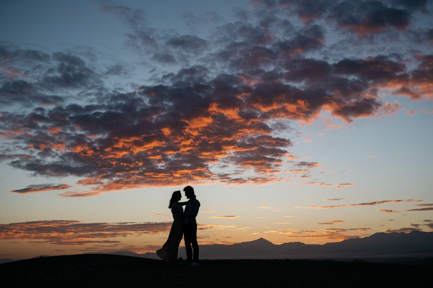 Silhouettes of a couple embrace against a dramatic sunset sky with orange and purple clouds.