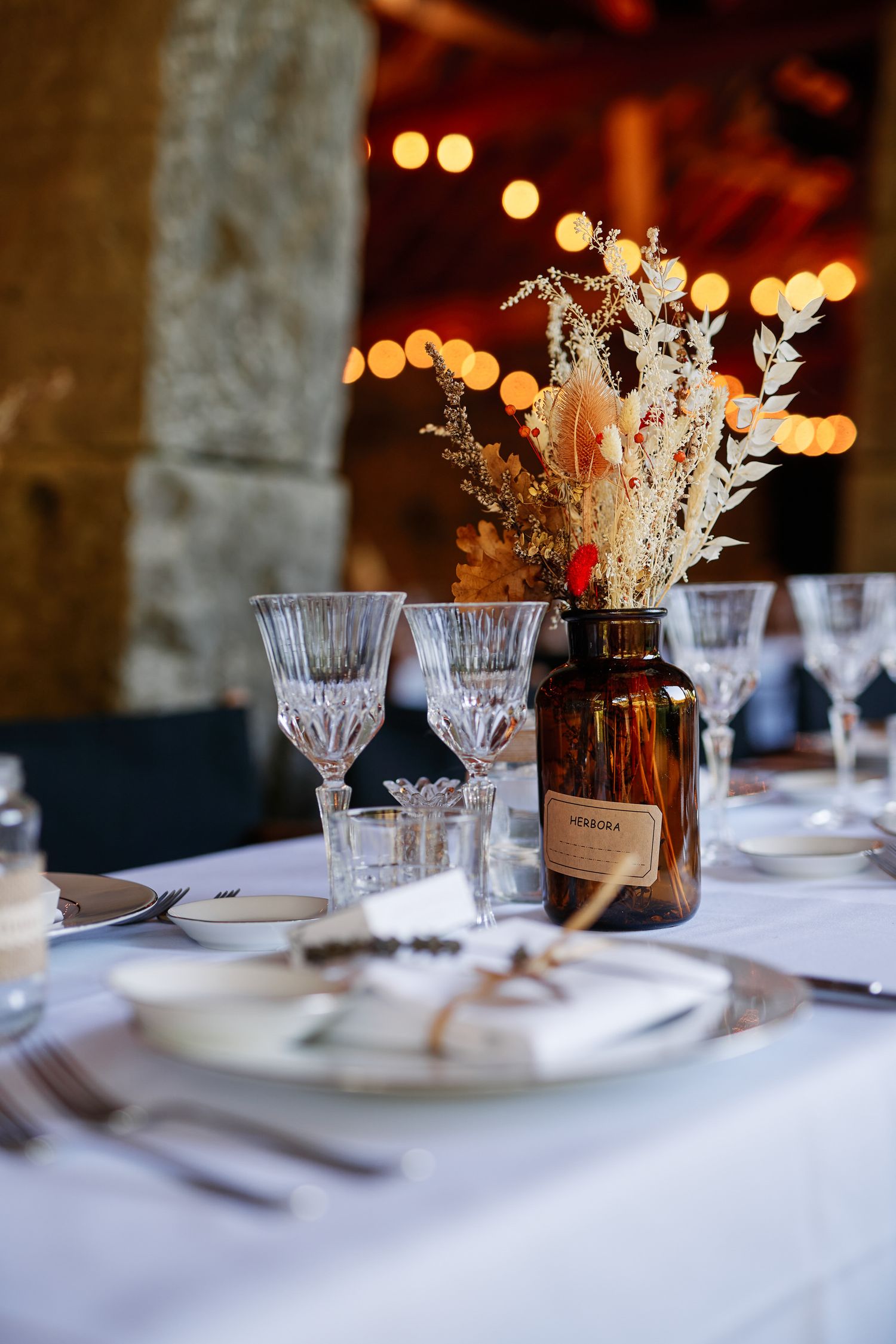 Elegant table setting with crystal glasses and dried flower centerpieces on white tablecloth with bokeh lights in background.
