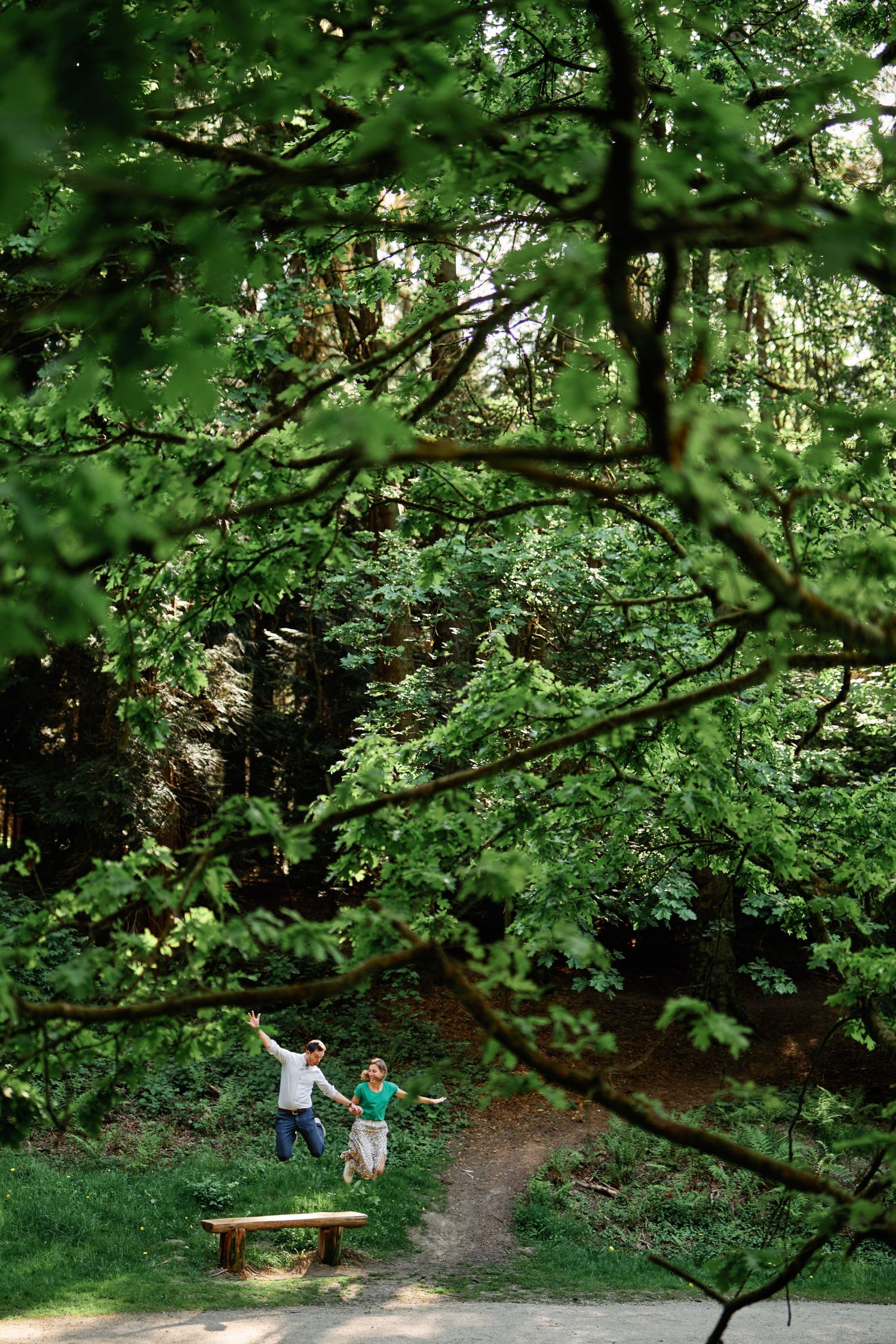 Two people walk along a shaded forest trail beneath green leafy tree branches on a sunny summer day.