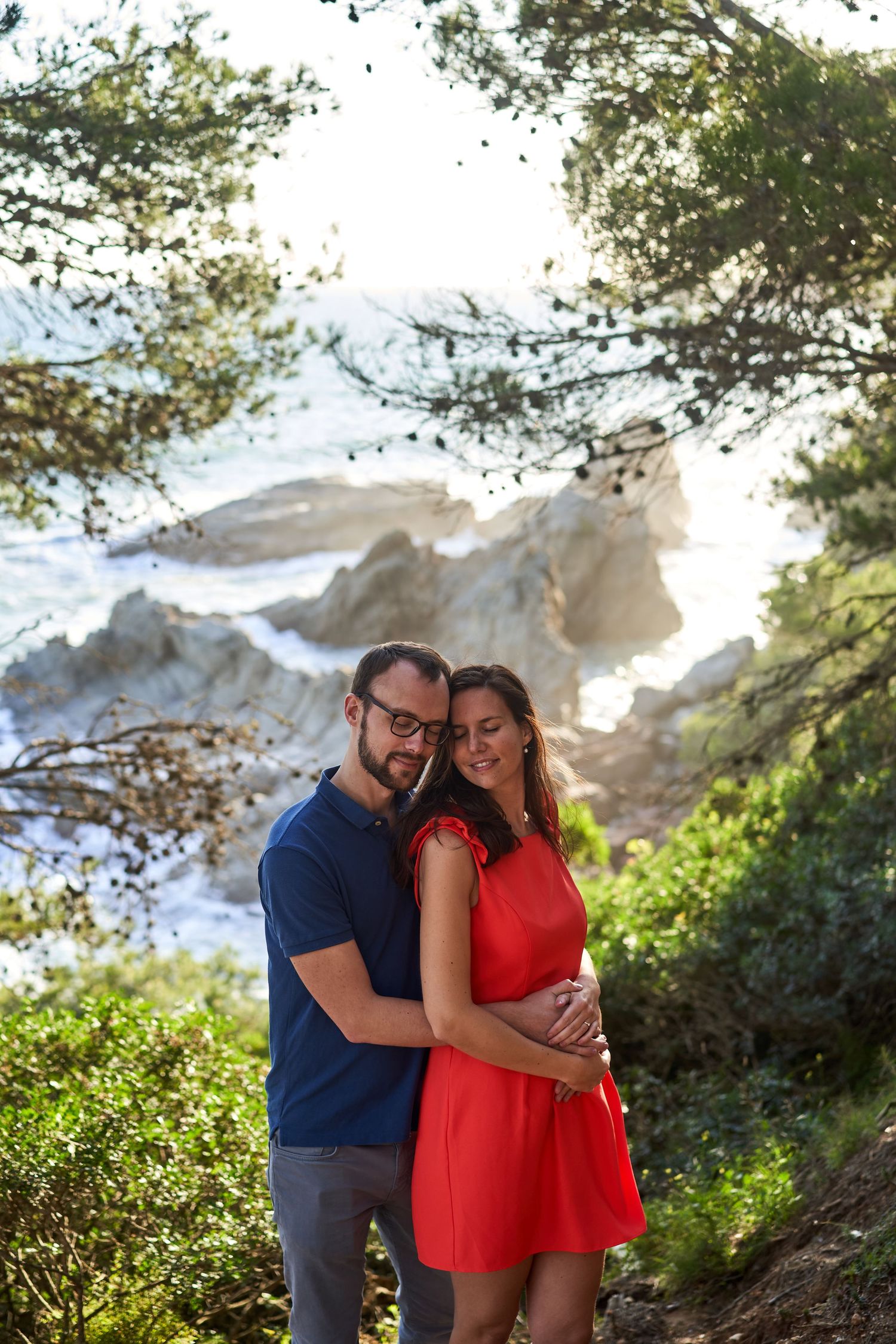 A couple embraces on a scenic mountain trail with rocky cliffs and greenery in the background.
