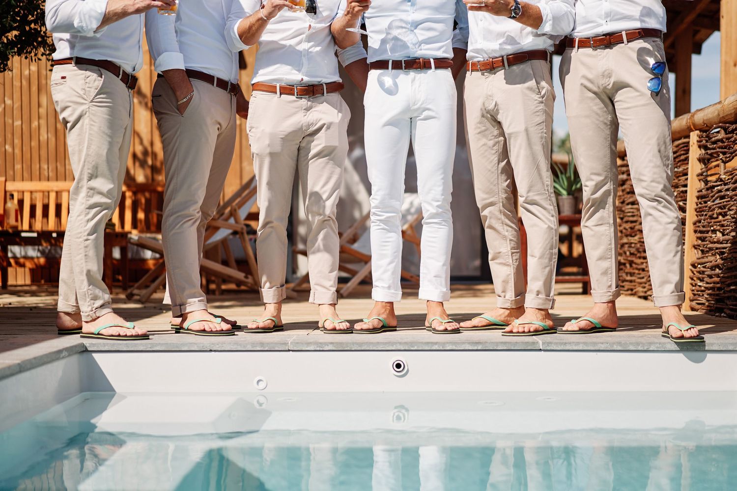 Wedding guests in white pants and beige shoes stand in a line along the edge of a swimming pool.