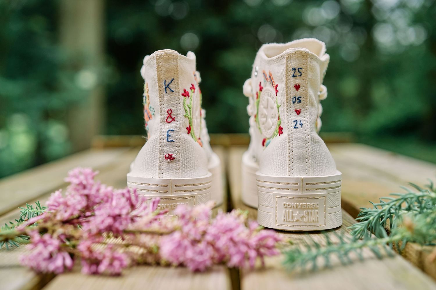 White Converse sneakers decorated with floral elements sit among pink flowers and greenery on a wooden surface.