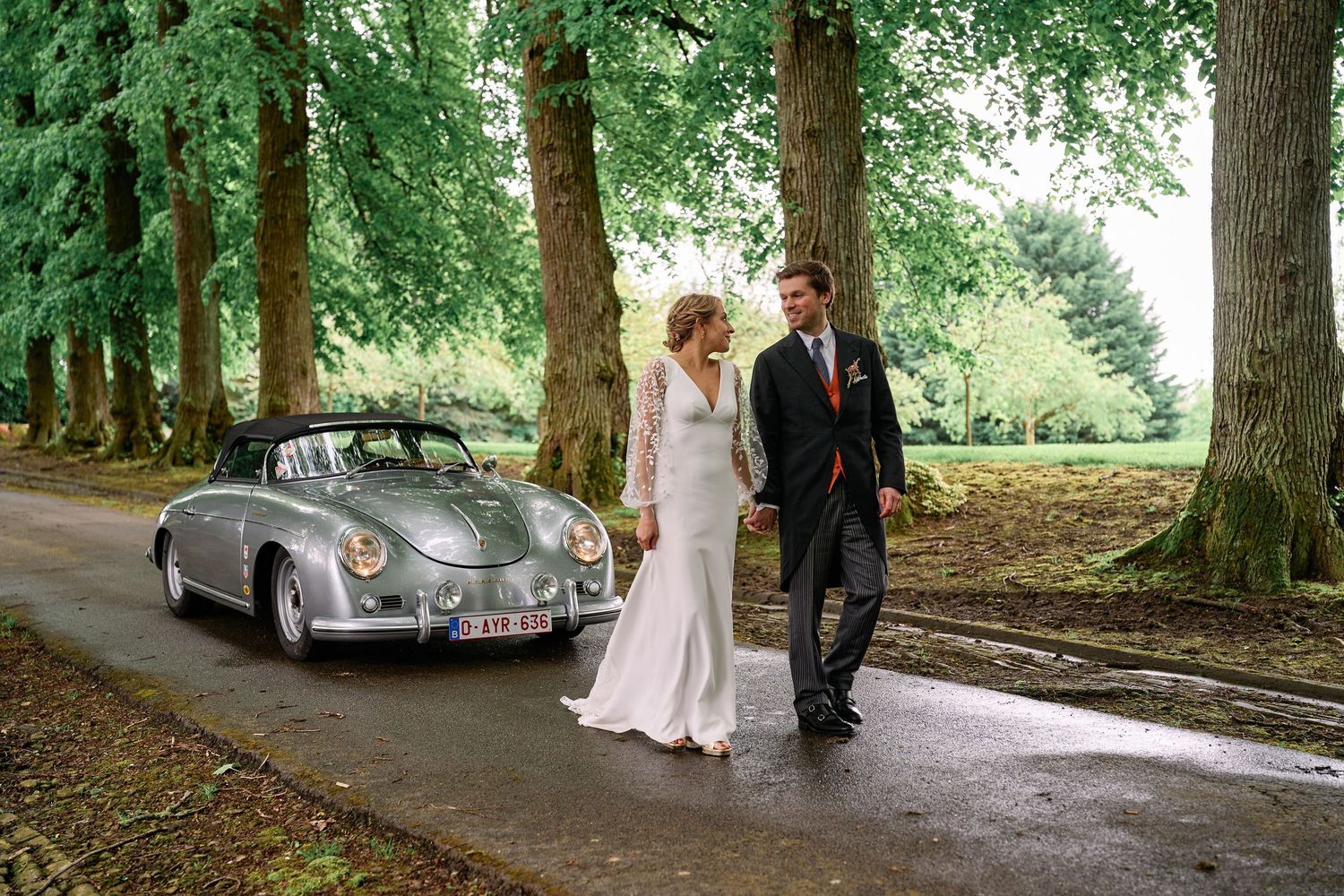 A classic vintage car parked under tall trees on a shaded path during a special occasion.