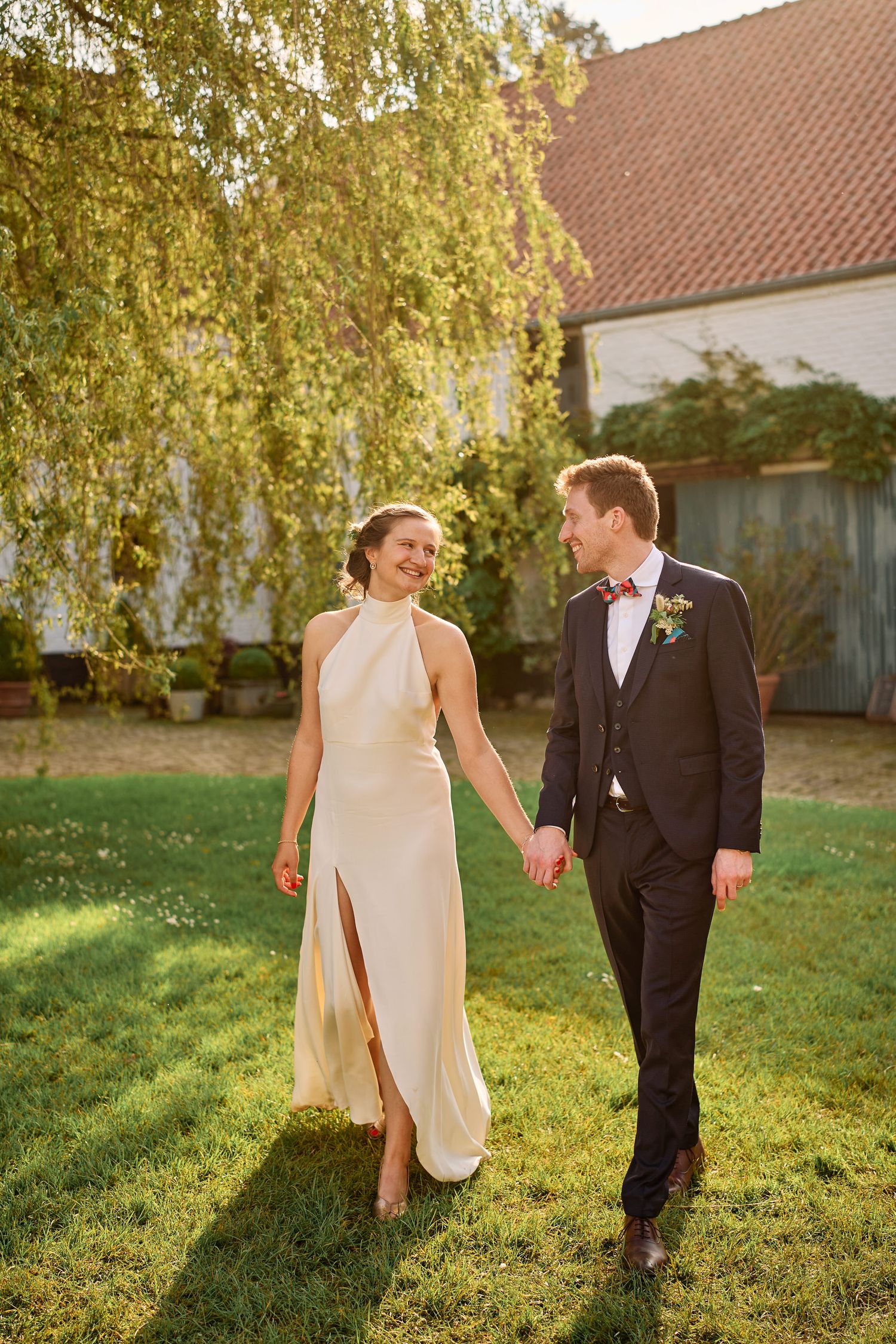 A couple in wedding attire walks through a sunlit garden during golden hour, creating a romantic outdoor scene.