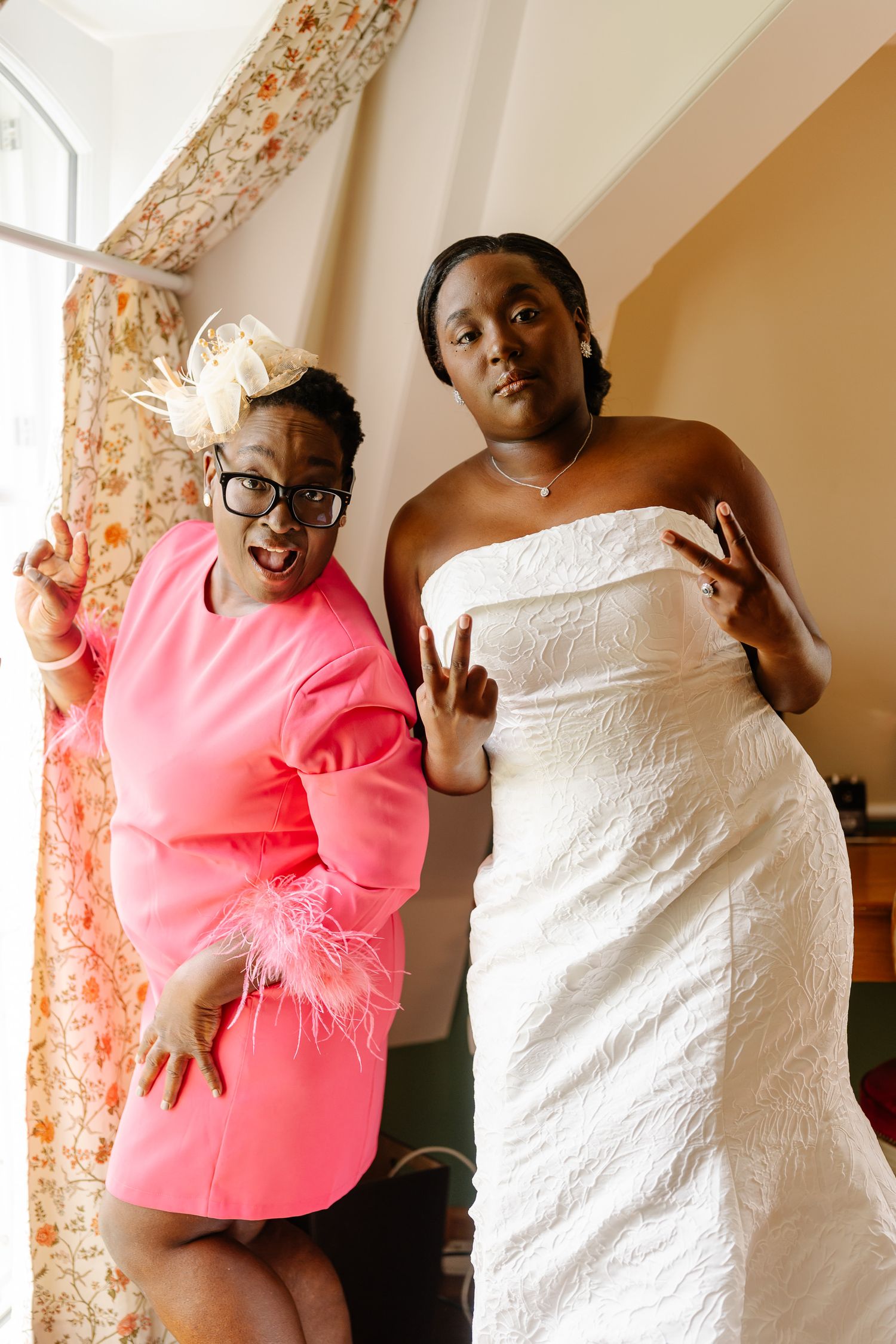 Two people pose together, one wearing a pink feathered dress and the other in a white strapless wedding gown.