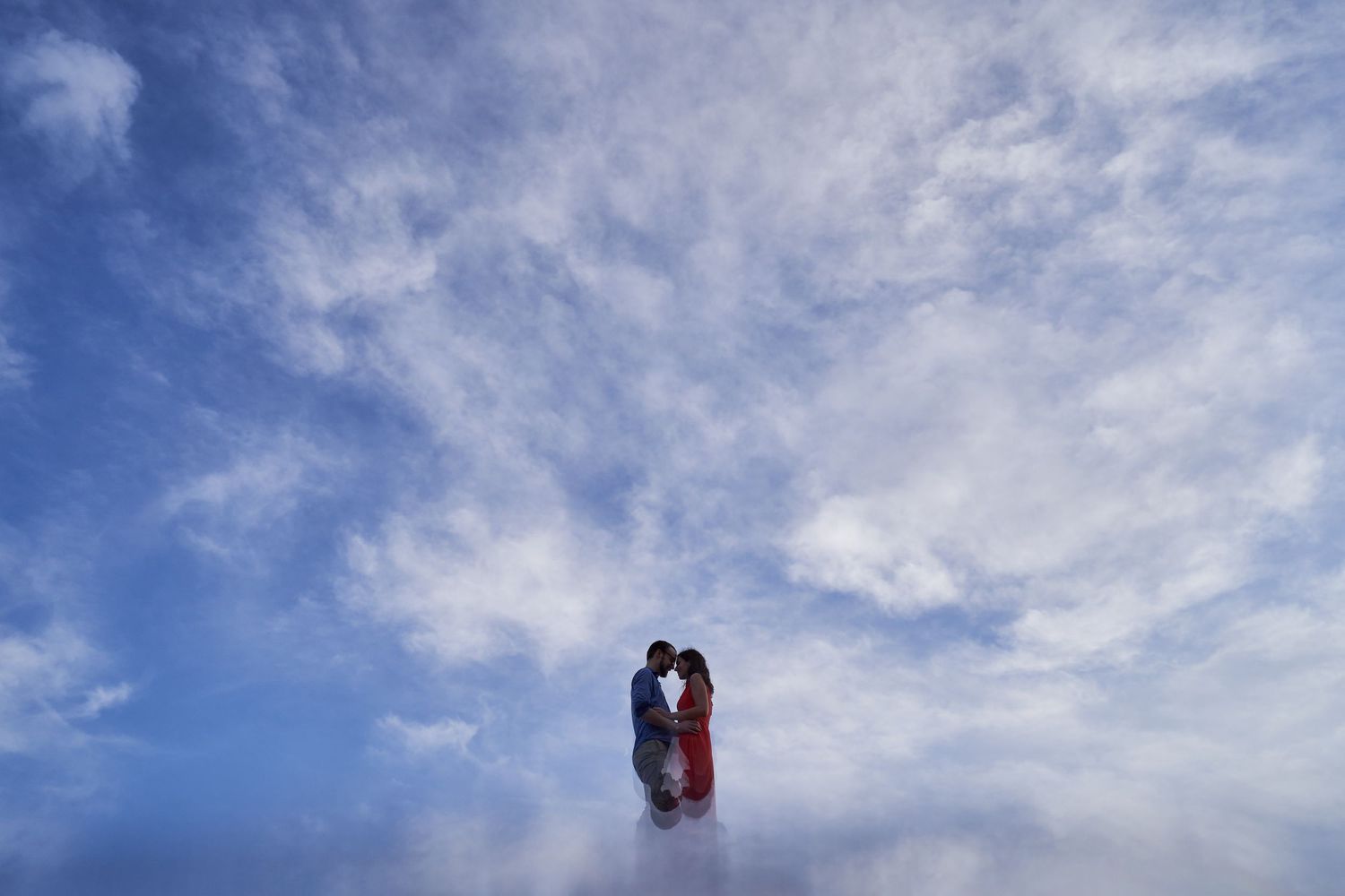 A silhouetted couple embraces against a dramatic blue sky with wispy white clouds.