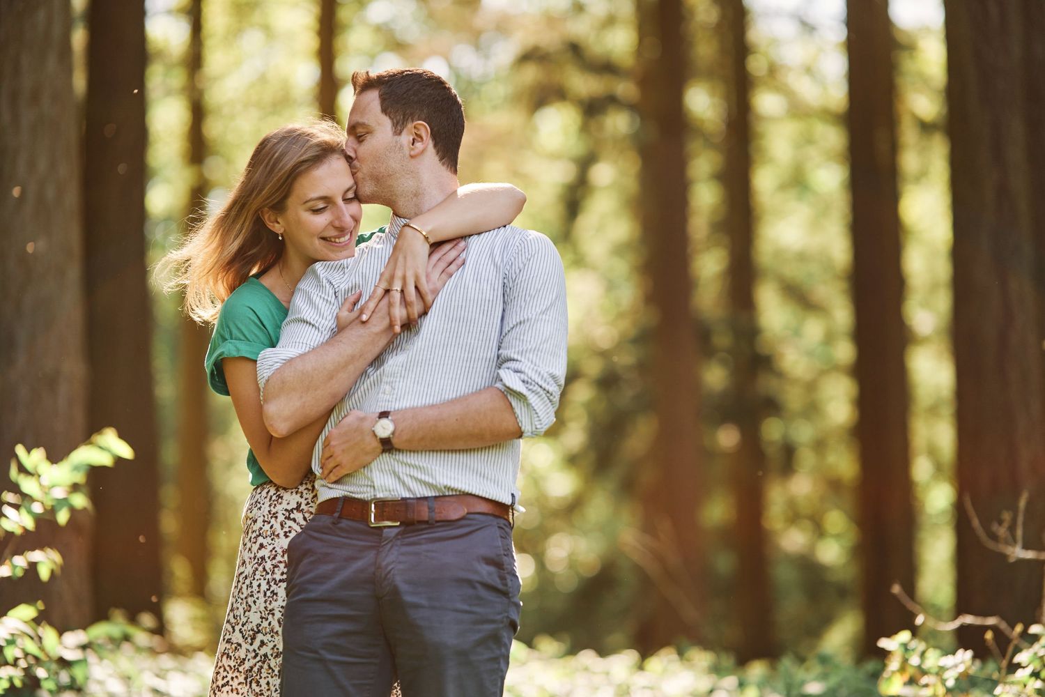 A couple embraces lovingly in a sunlit forest setting during a romantic engagement photo session.