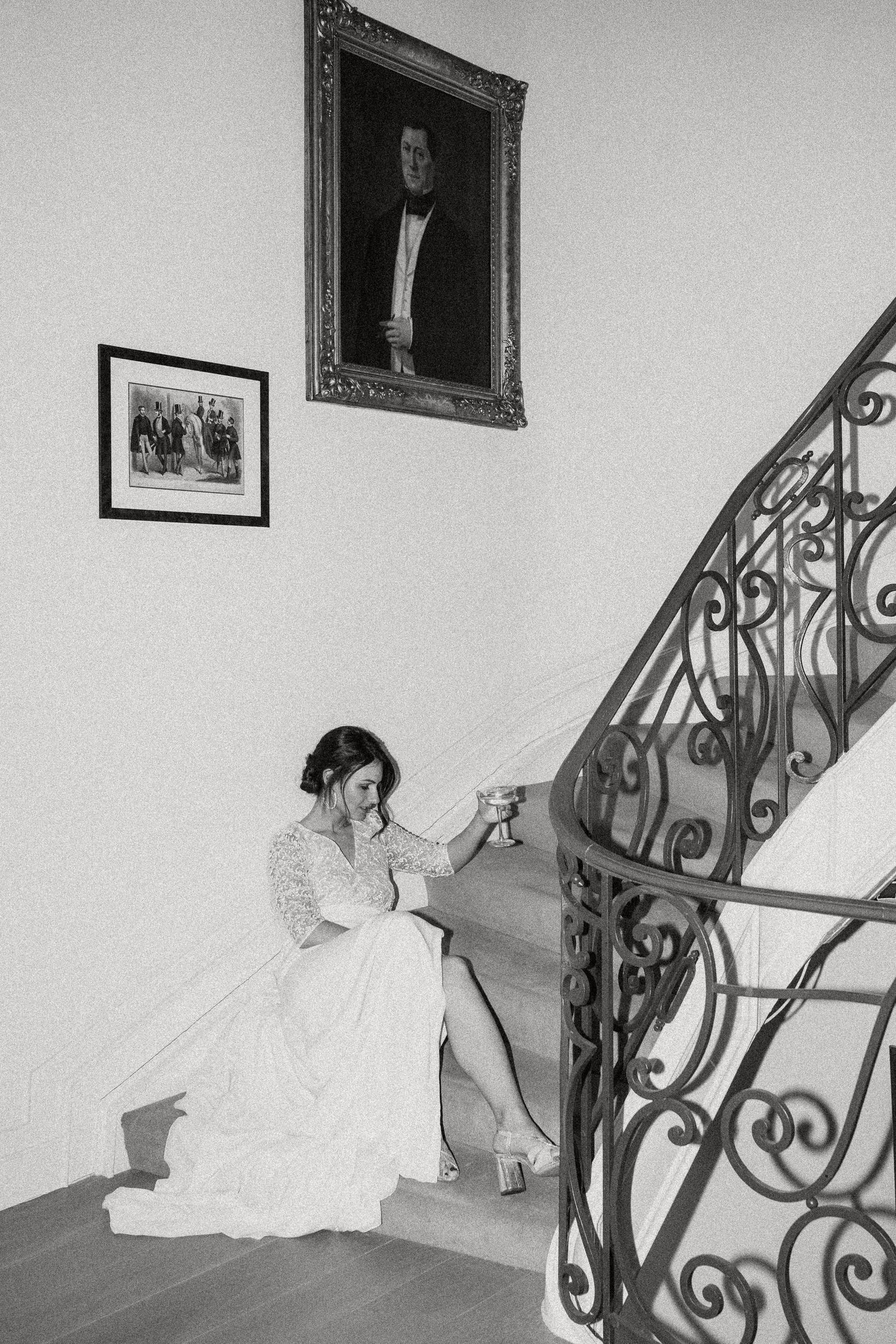 A black and white photo showing a figure in a white dress sitting on ornate staircase steps beneath a framed portrait.