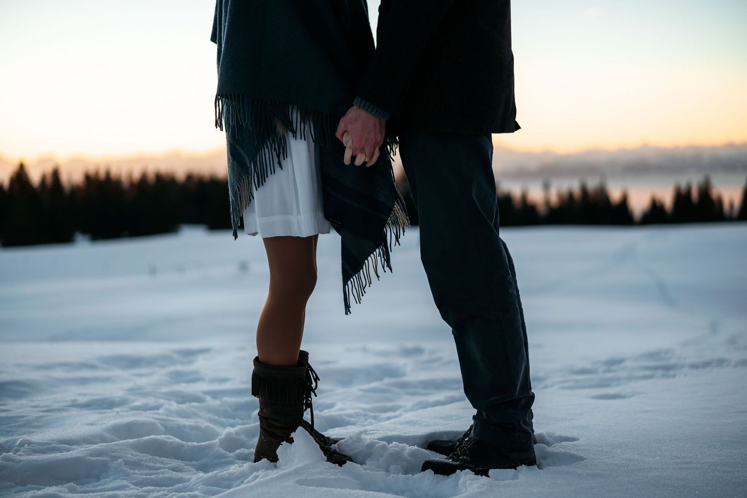 Two people stand close together on a frozen lake at dusk with snow-covered trees in the background.