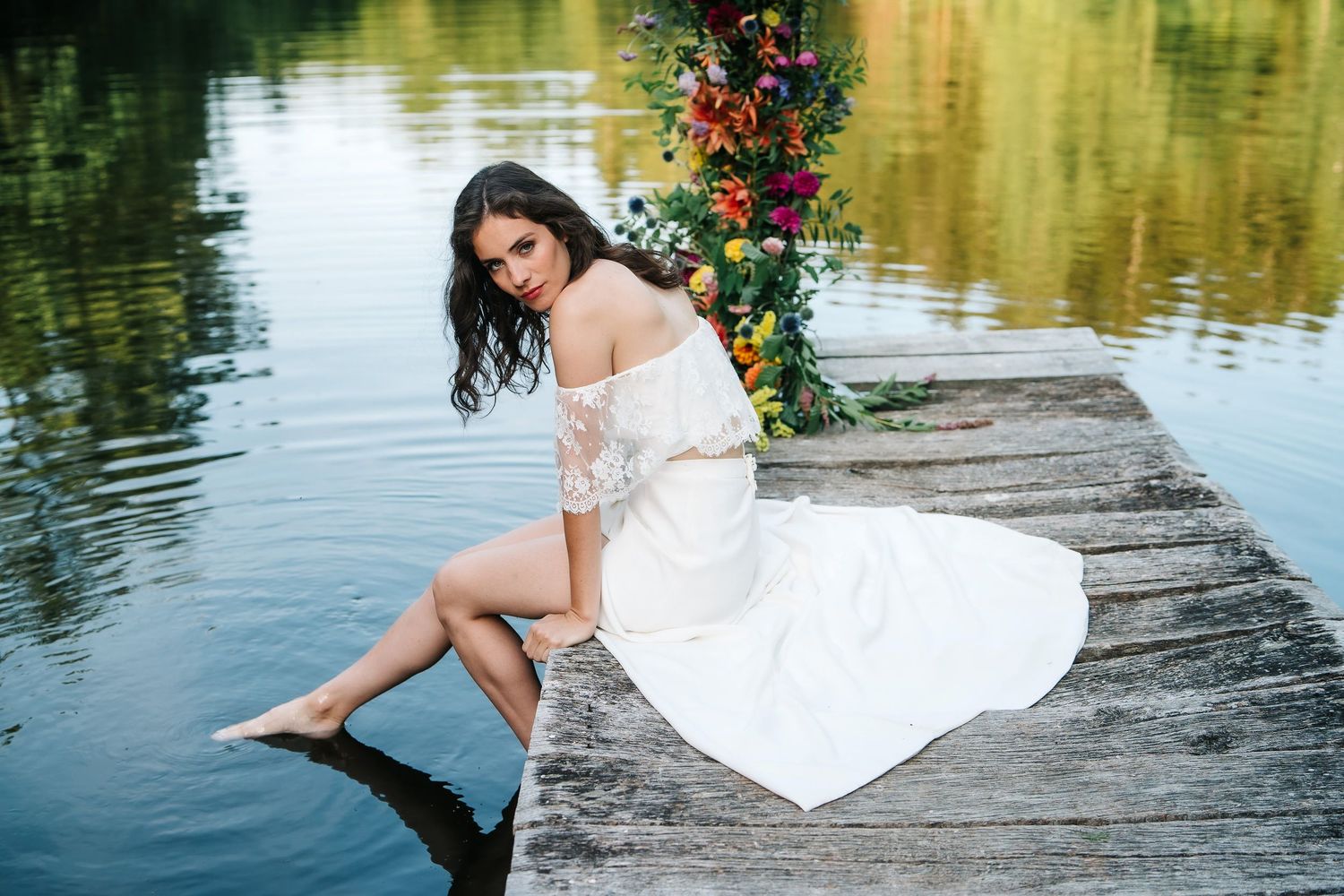 A woman in a flowing white dress poses with a husky dog near a lake surrounded by evergreen trees.