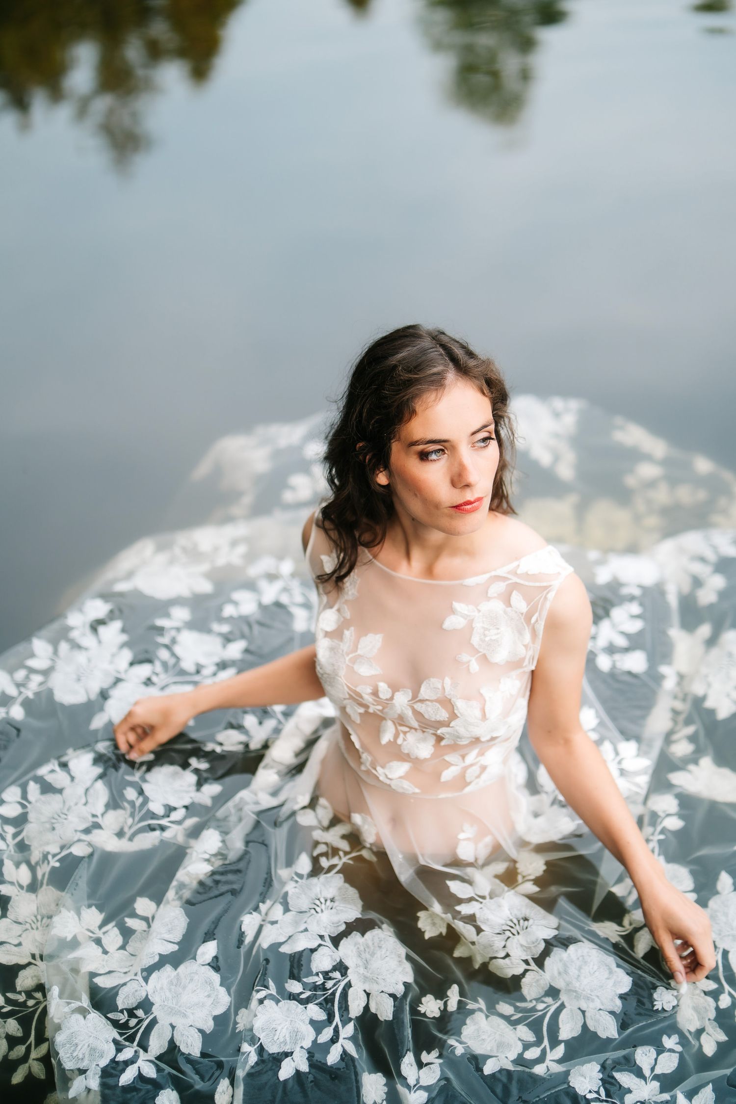A dreamy portrait of someone in a white floral dress floating in dark reflective water with scattered white flowers.