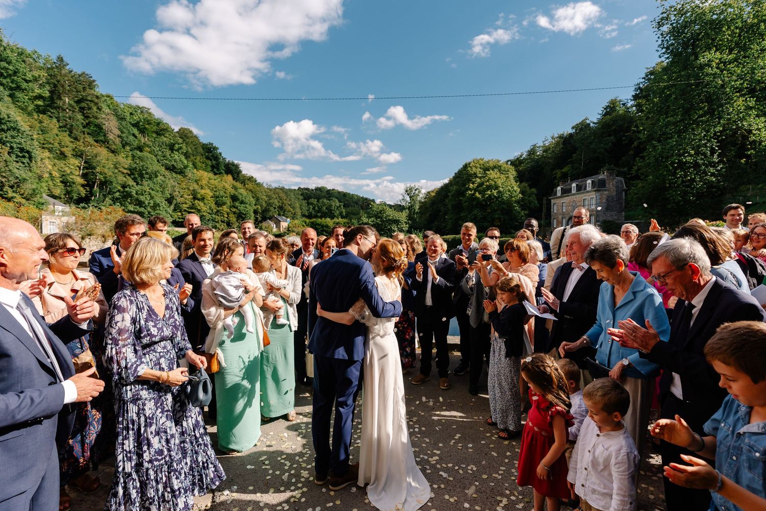 Outdoor wedding celebration with guests gathered on a sunny day surrounded by lush green trees and blue skies.