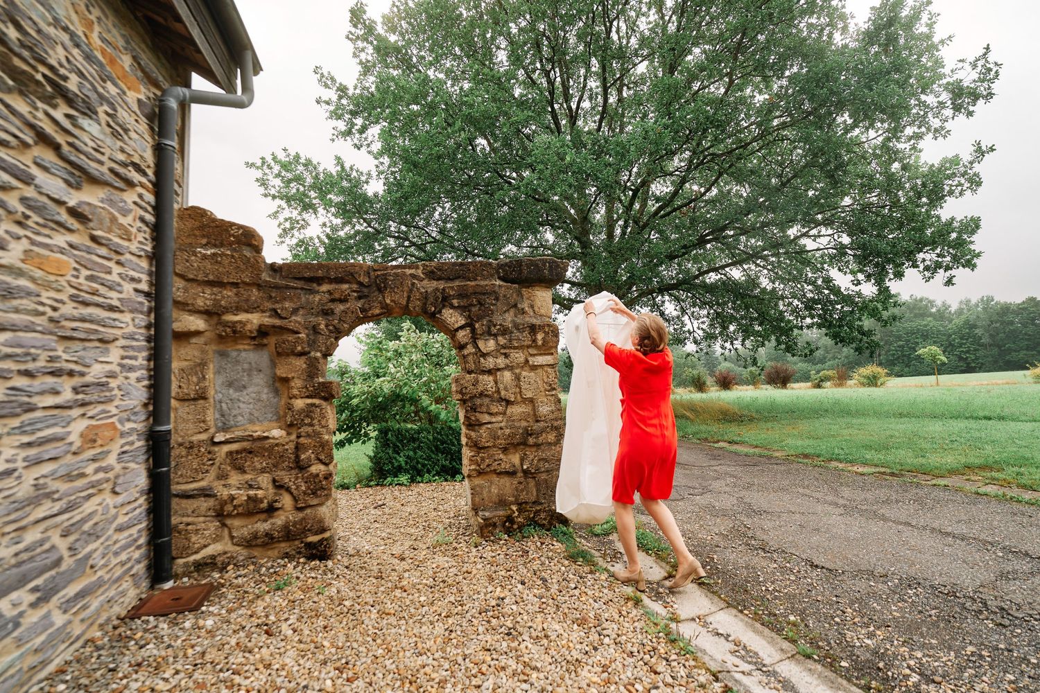 A person in a bright red dress poses playfully against an old stone wall with a green tree in the background.