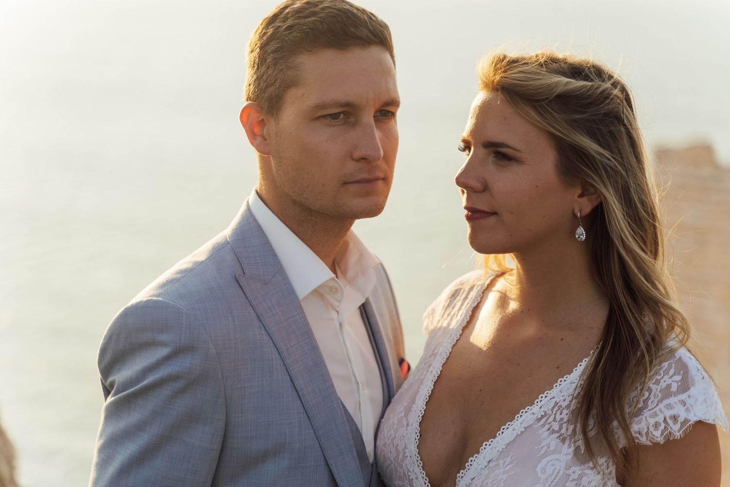 A couple in wedding attire share an intimate moment at sunset, with the bride in lace and groom in light gray suit.