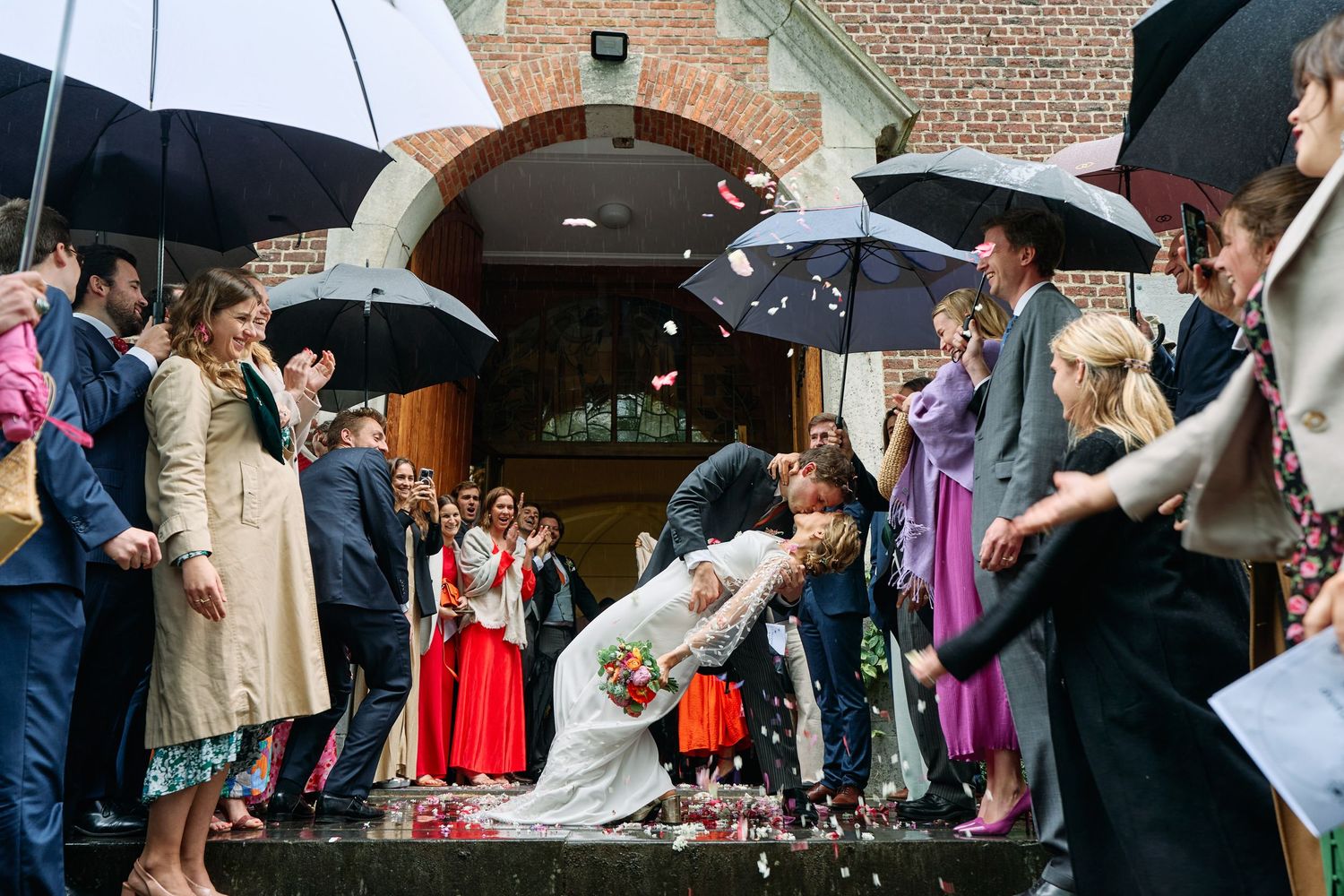 Wedding guests hold umbrellas and celebrate as newlyweds exit church on rainy day.