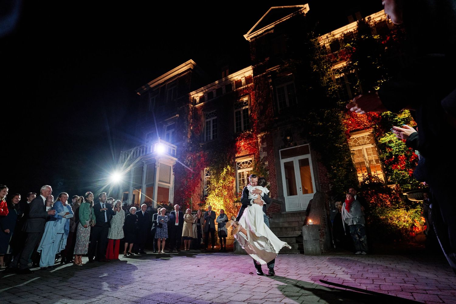 Outdoor nighttime wedding celebration with guests dancing under string lights and colorful illumination in a garden setting.