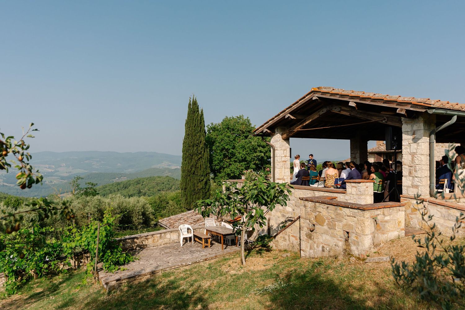 Panoramic view of rustic stone buildings and rolling green hills in the Tuscan countryside on a sunny day.
