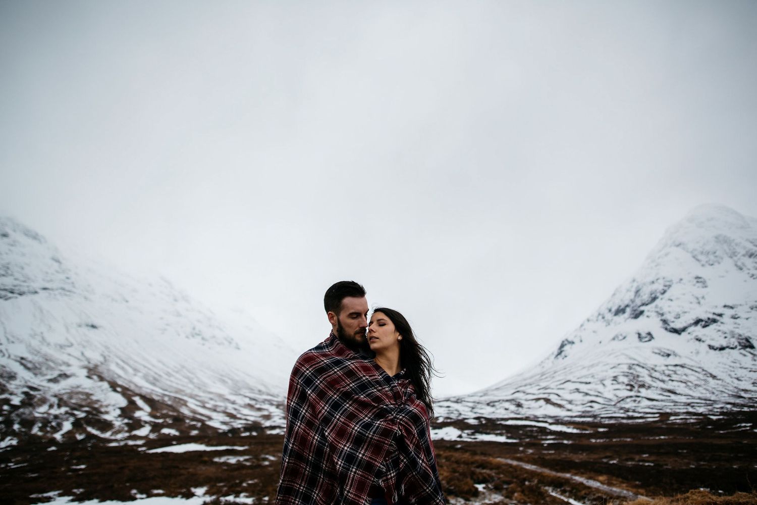 A couple embraces against a dramatic backdrop of snow-capped mountains in Scotland's rugged wilderness.