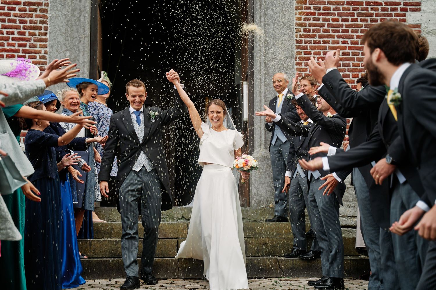 Wedding party celebrates by throwing rice and confetti at newlyweds outside brick church building.