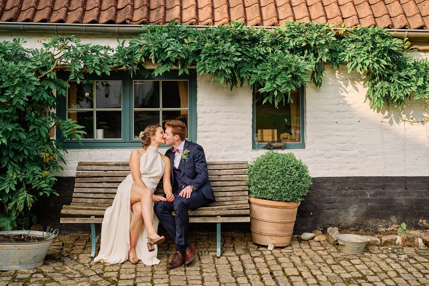 A couple shares an intimate moment on a rustic wooden bench outside a charming farmhouse with terracotta roof tiles.
