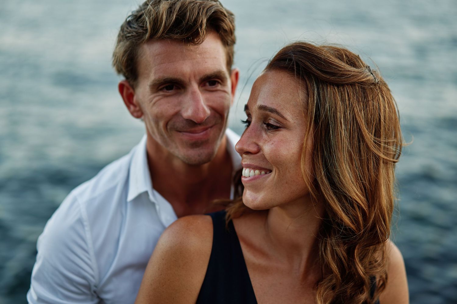Romantic couple embraces and smiles while sitting by the ocean at sunset with waves in background.