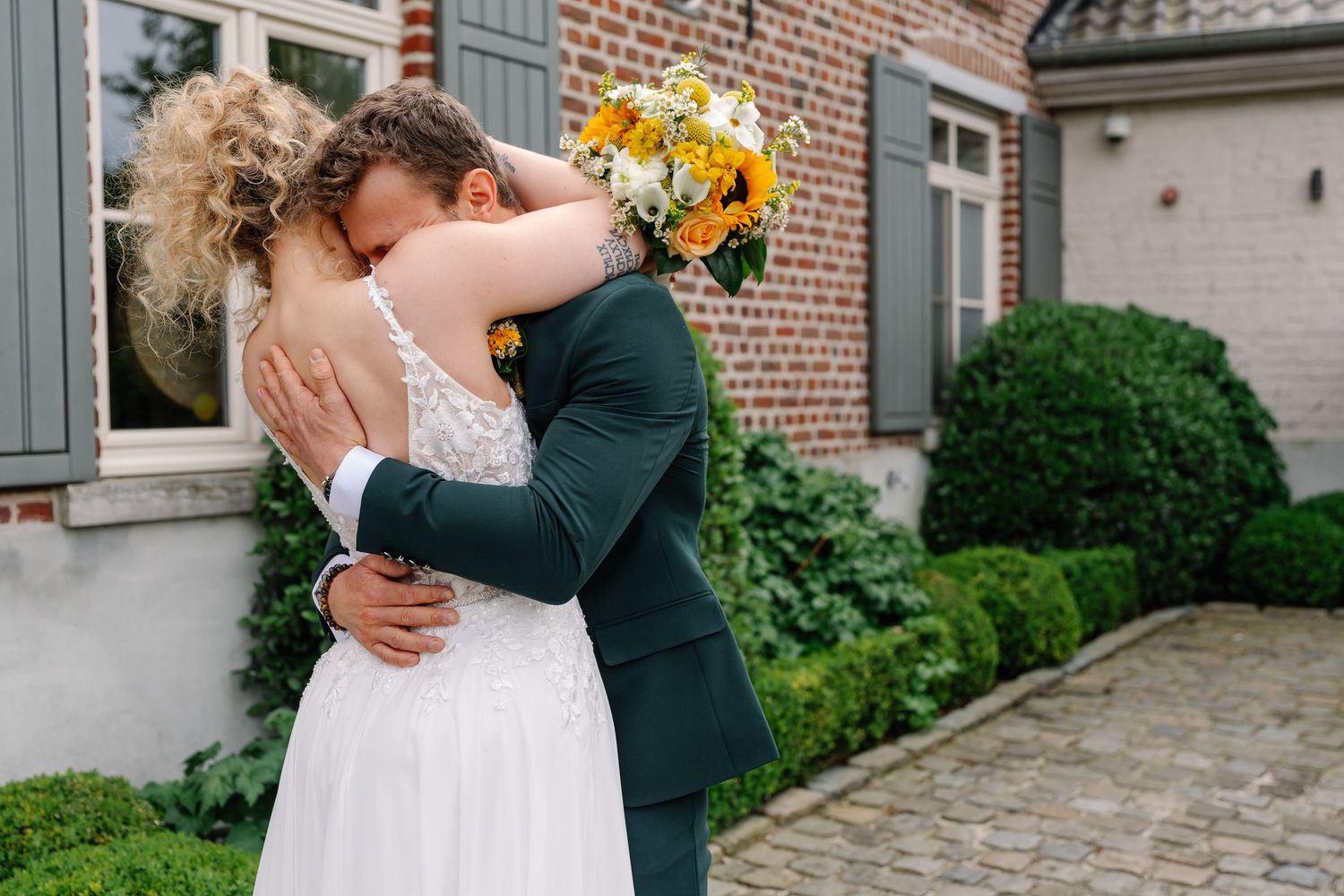 A romantic couple embraces during their wedding day in front of a rustic brick building with lush greenery.