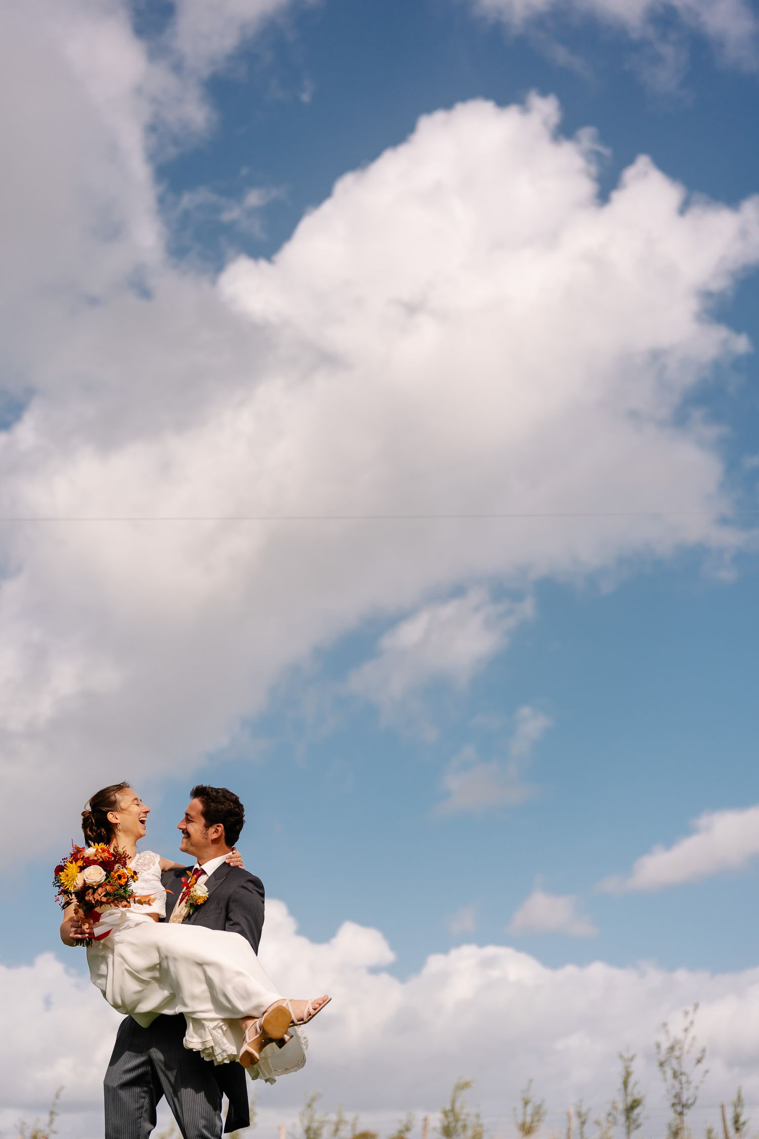 A couple embraces joyfully against a bright blue sky with white fluffy clouds on their wedding day.