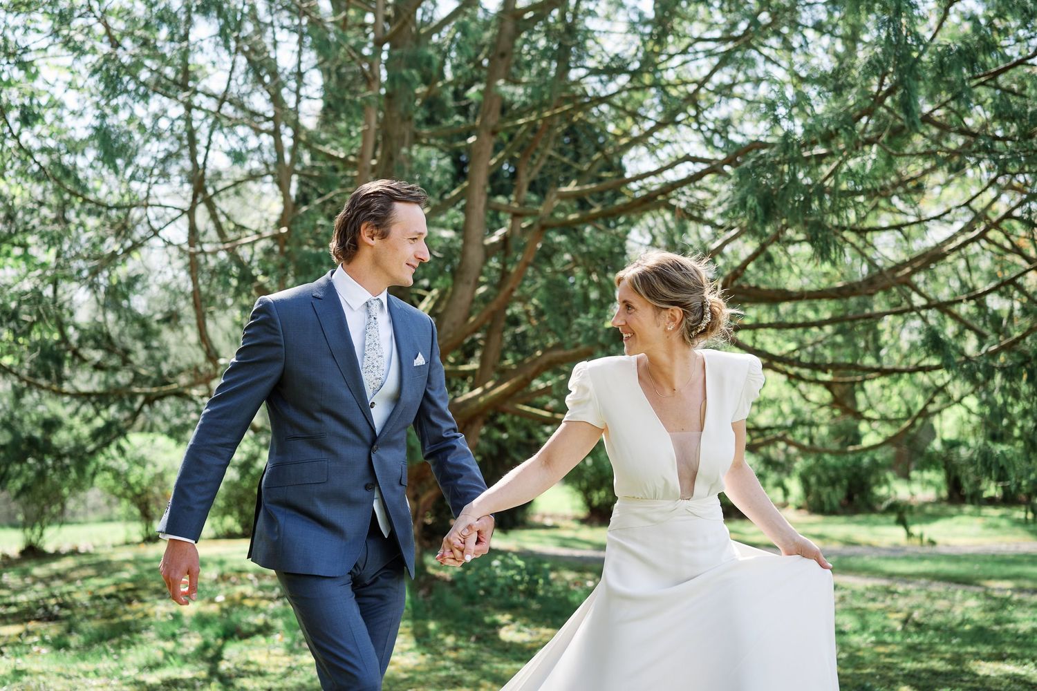 A couple in wedding attire dances together beneath towering evergreen trees in a sunlit forest setting.
