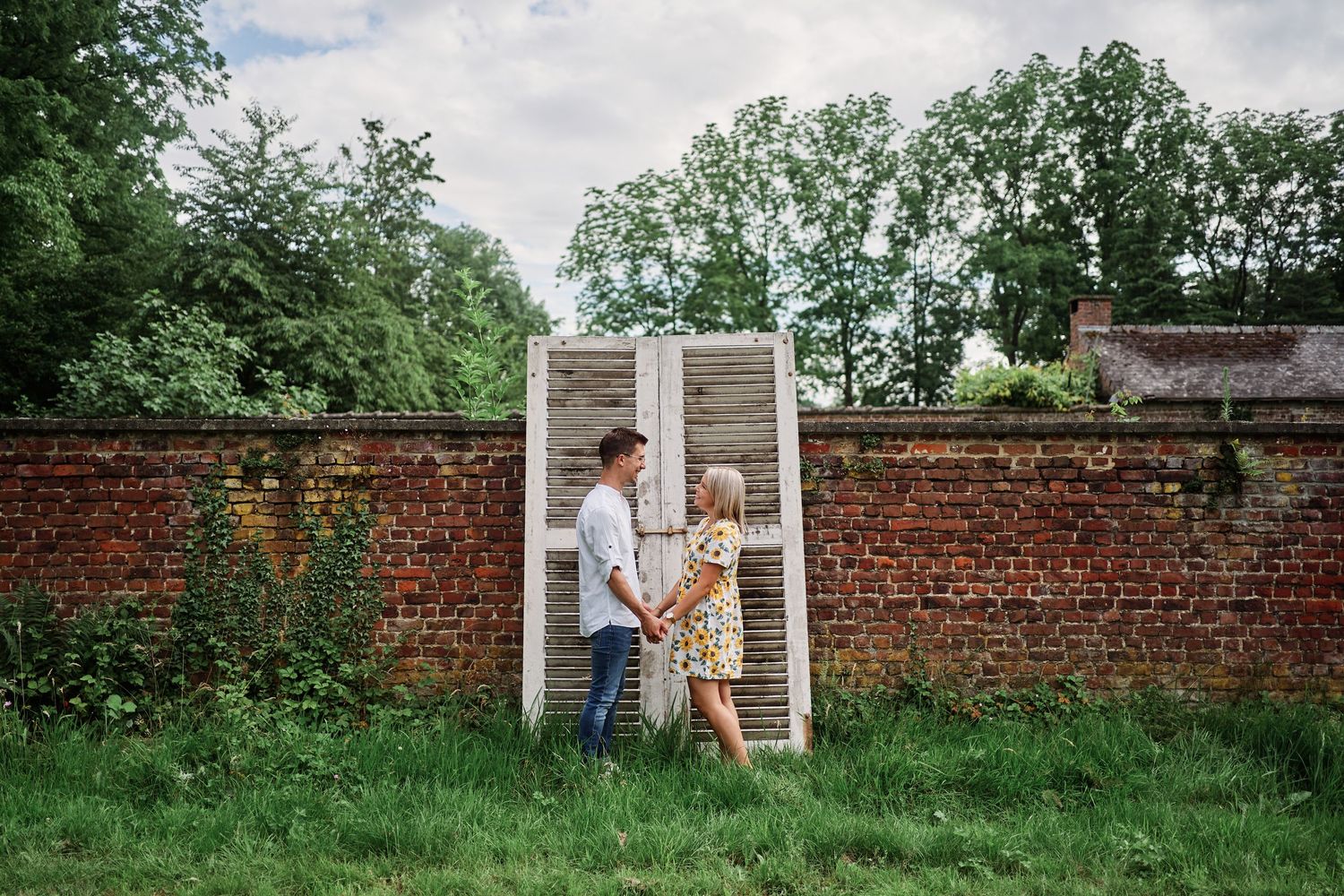 Couple in casual attire poses playfully against a rustic brick wall in a garden setting.