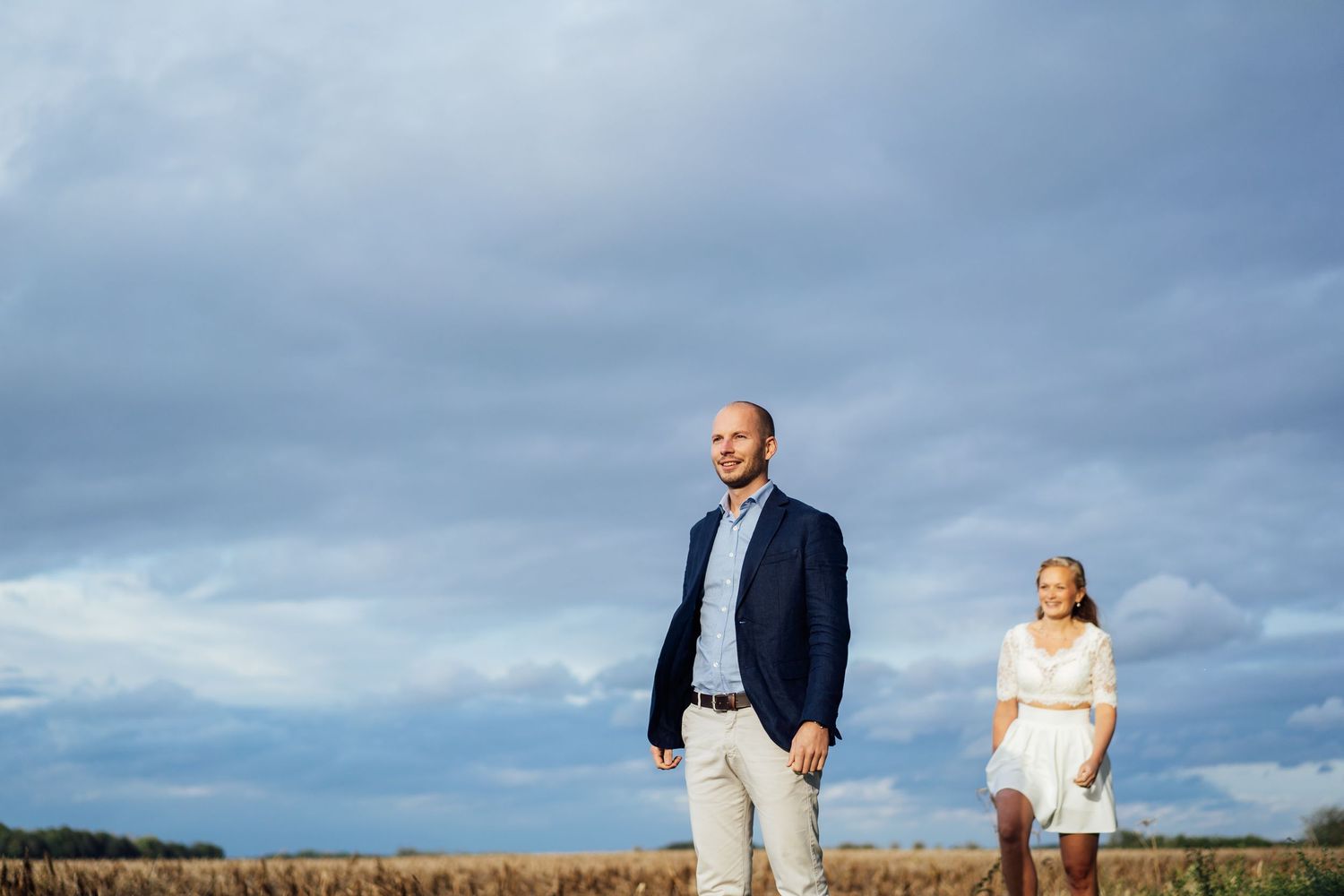 Couple walking together through a scenic field at sunset against dramatic cloudy skies.