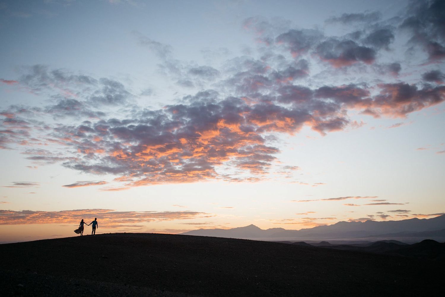 Two silhouettes walk along a ridge against a dramatic sunset sky with pink and orange clouds.
