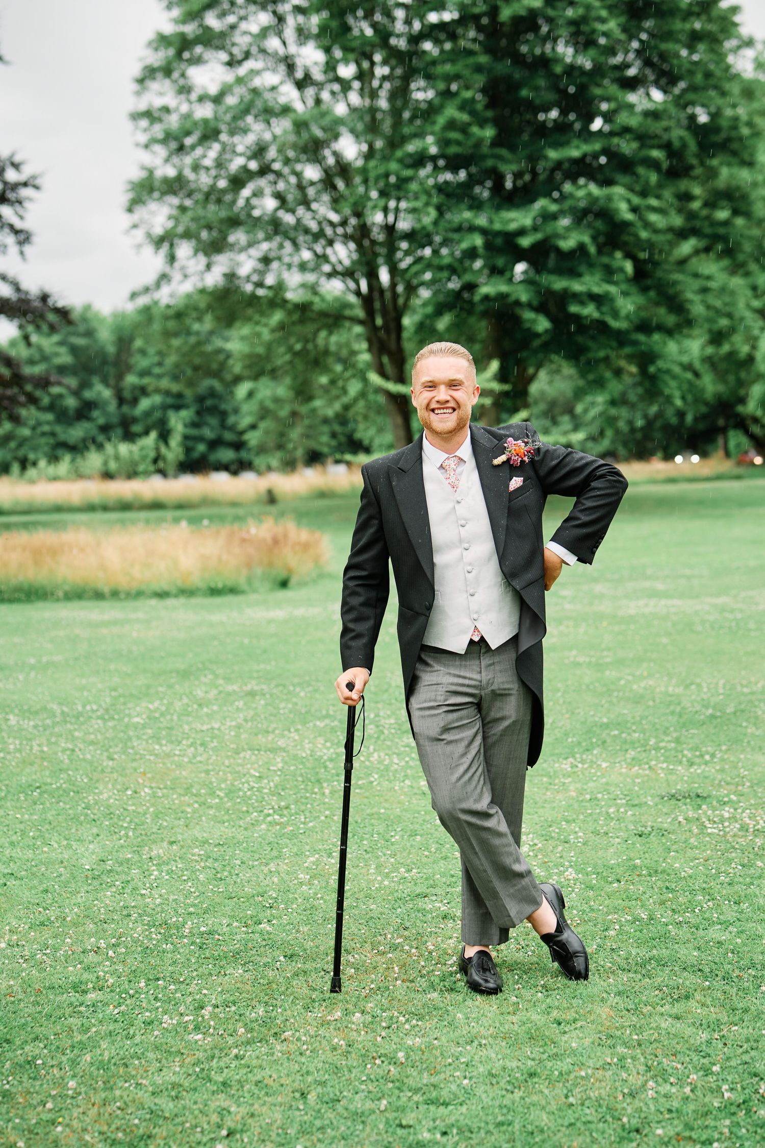 A person in a formal tuxedo walks with a cane across a green lawn dotted with white flowers on a sunny day.