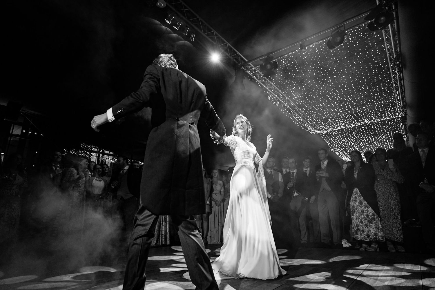 A wedding couple dances under twinkling lights during their first dance at an evening reception.