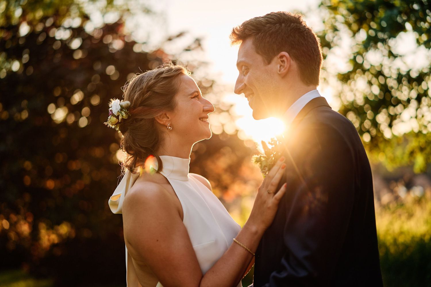 Newlywed couple embraces during golden hour sunset with sunlight streaming through trees in outdoor wedding setting.