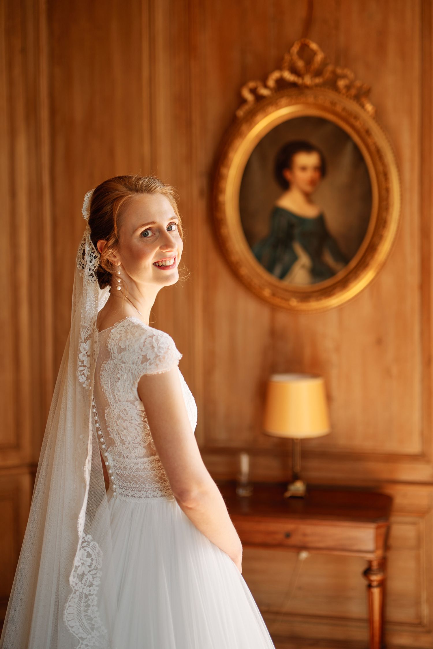Bride in lace cap sleeve wedding dress and veil poses in front of vintage portrait and wooden wall.
