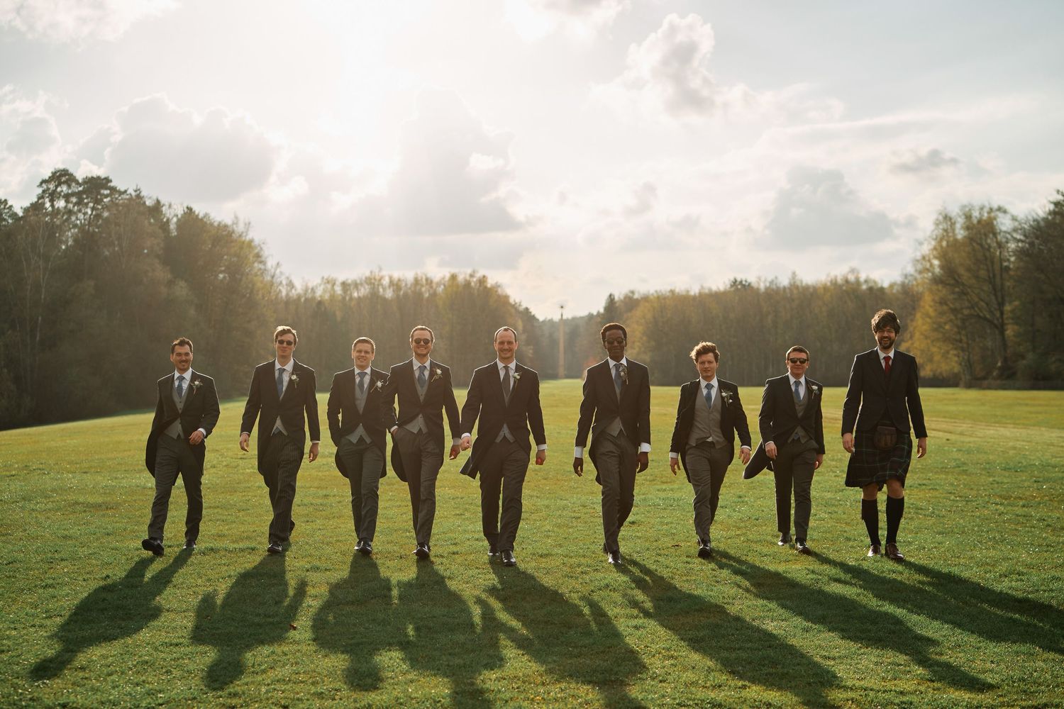 Groomsmen walking in a line across a sunlit field casting long shadows at sunset.