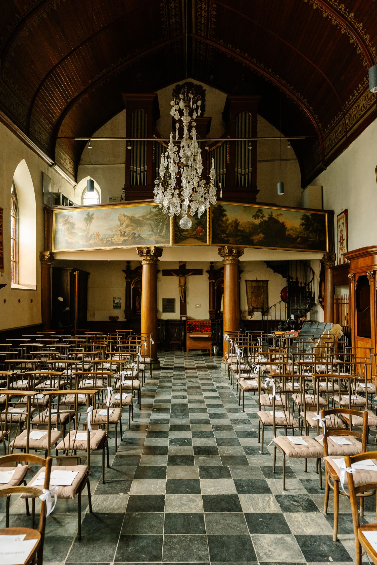 Interior of an elegant church venue with black and white checkered floor and rows of wooden chairs under crystal chandeliers.