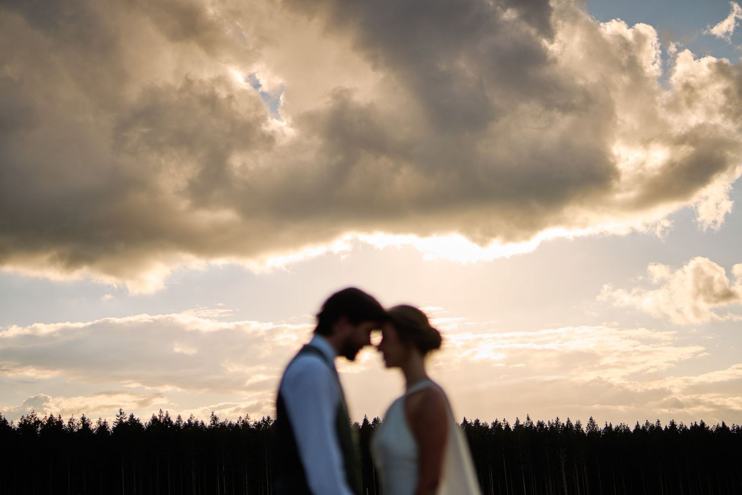 A dramatic sunset silhouettes a couple sharing a romantic kiss against a cloudy sky and forest backdrop.