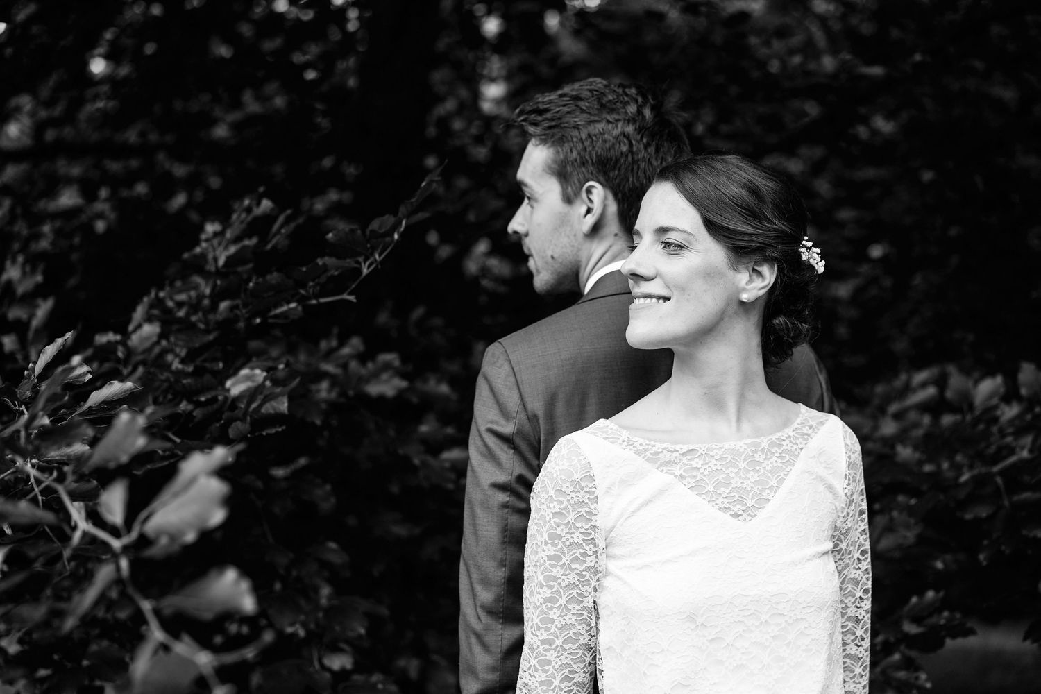 A black and white wedding photo showing a couple in formal attire standing together outdoors with natural foliage behind them.