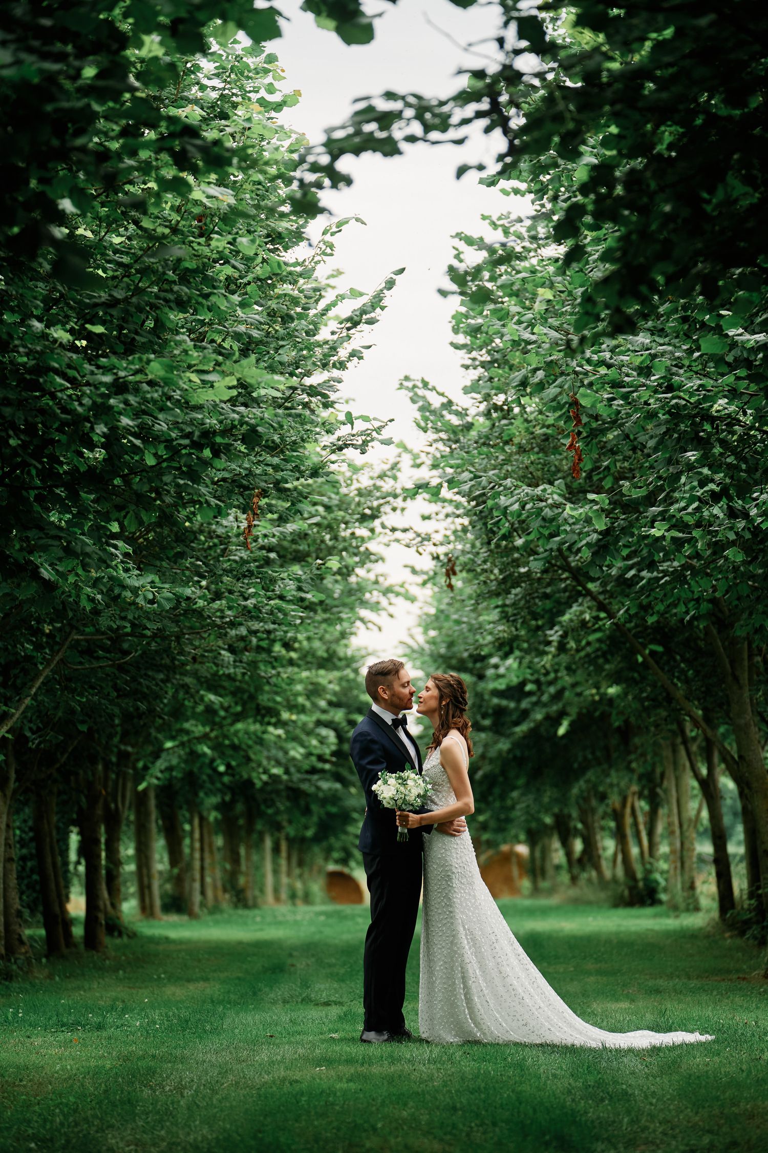 Newlyweds share romantic moments in a lush green tree-lined path during their outdoor wedding photoshoot.