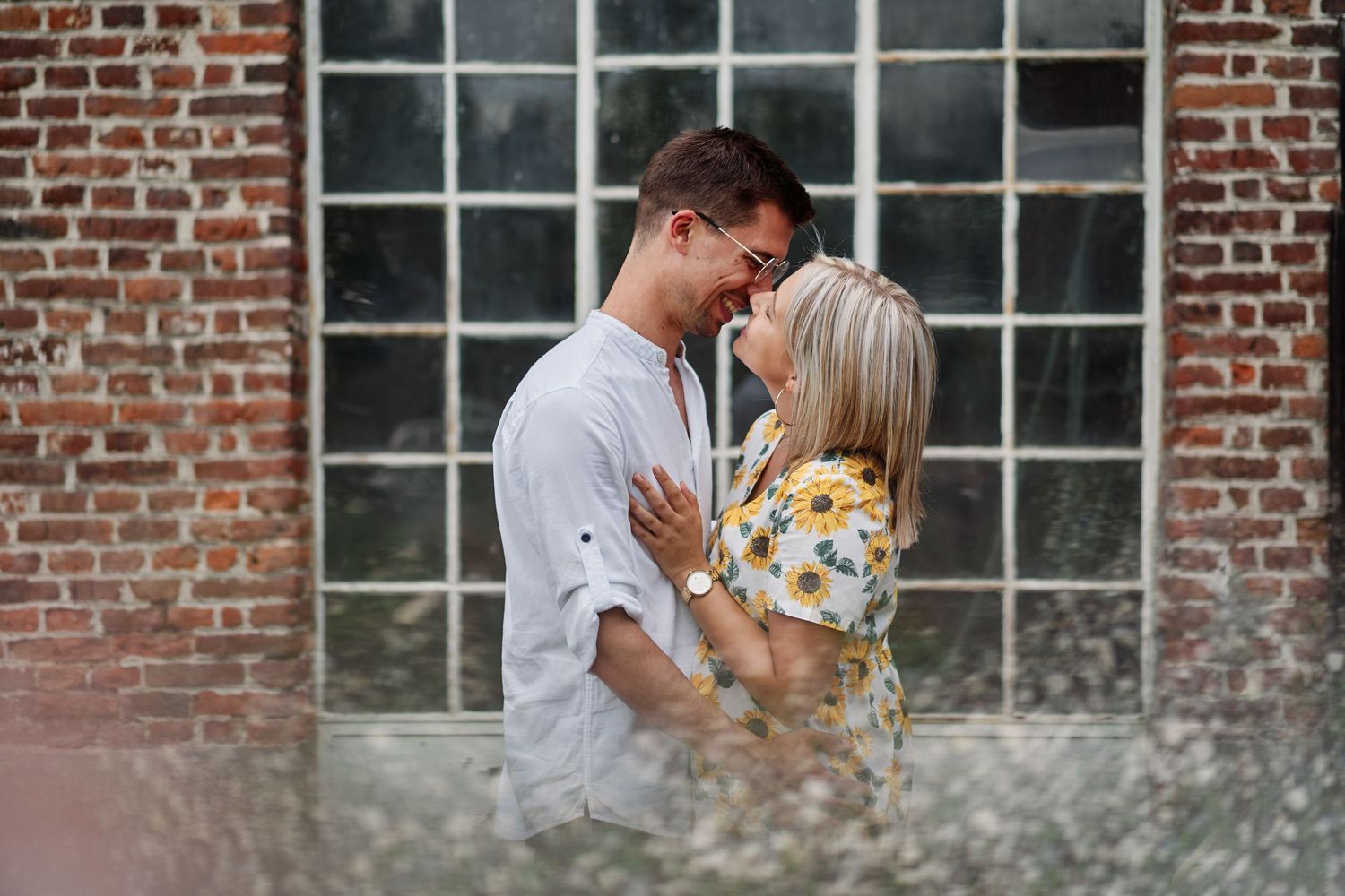 A romantic couple shares tender kisses against a rustic brick wall backdrop during an outdoor photo session.