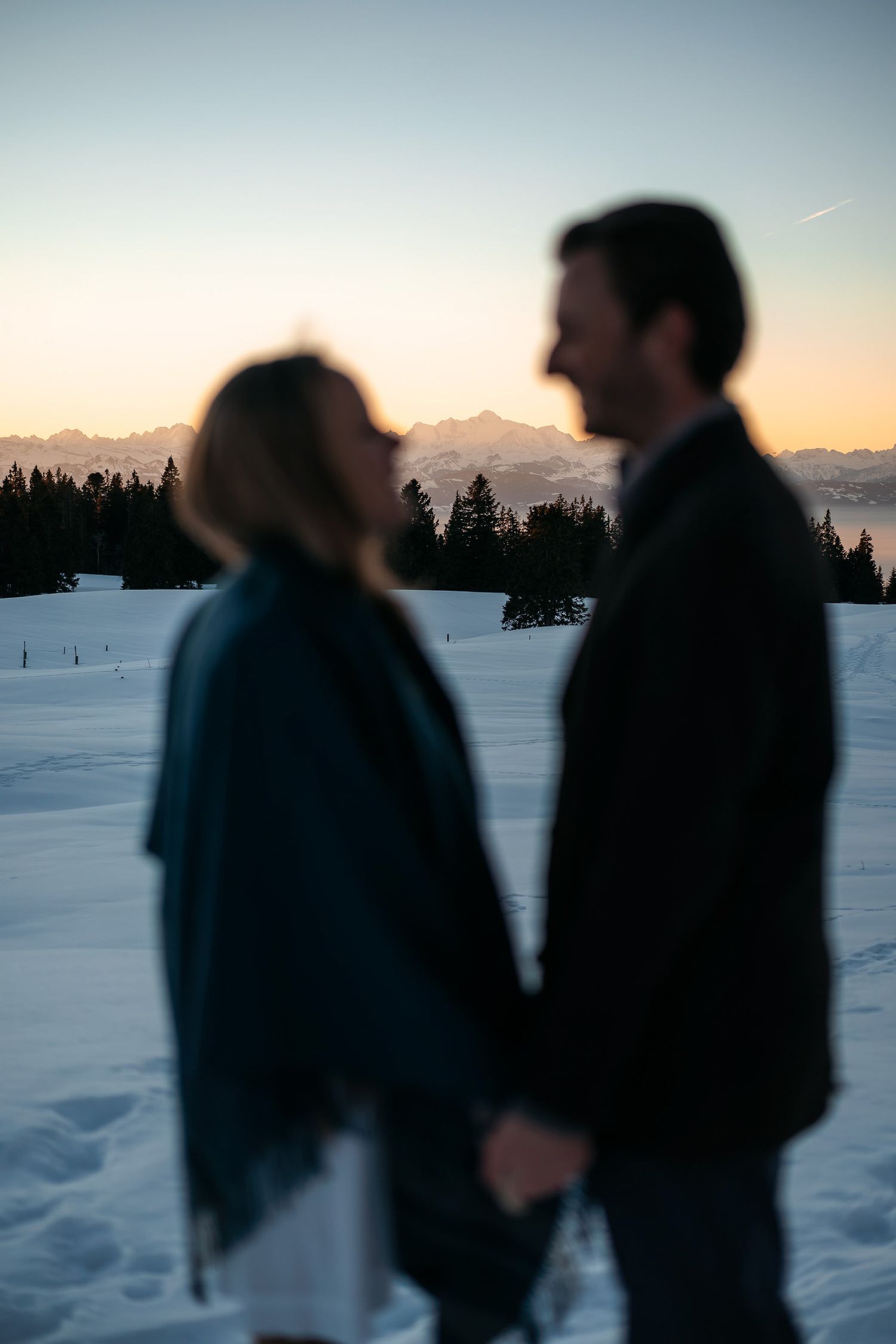 Silhouettes of people standing in a snowy winter landscape at sunset with mountains in the background.