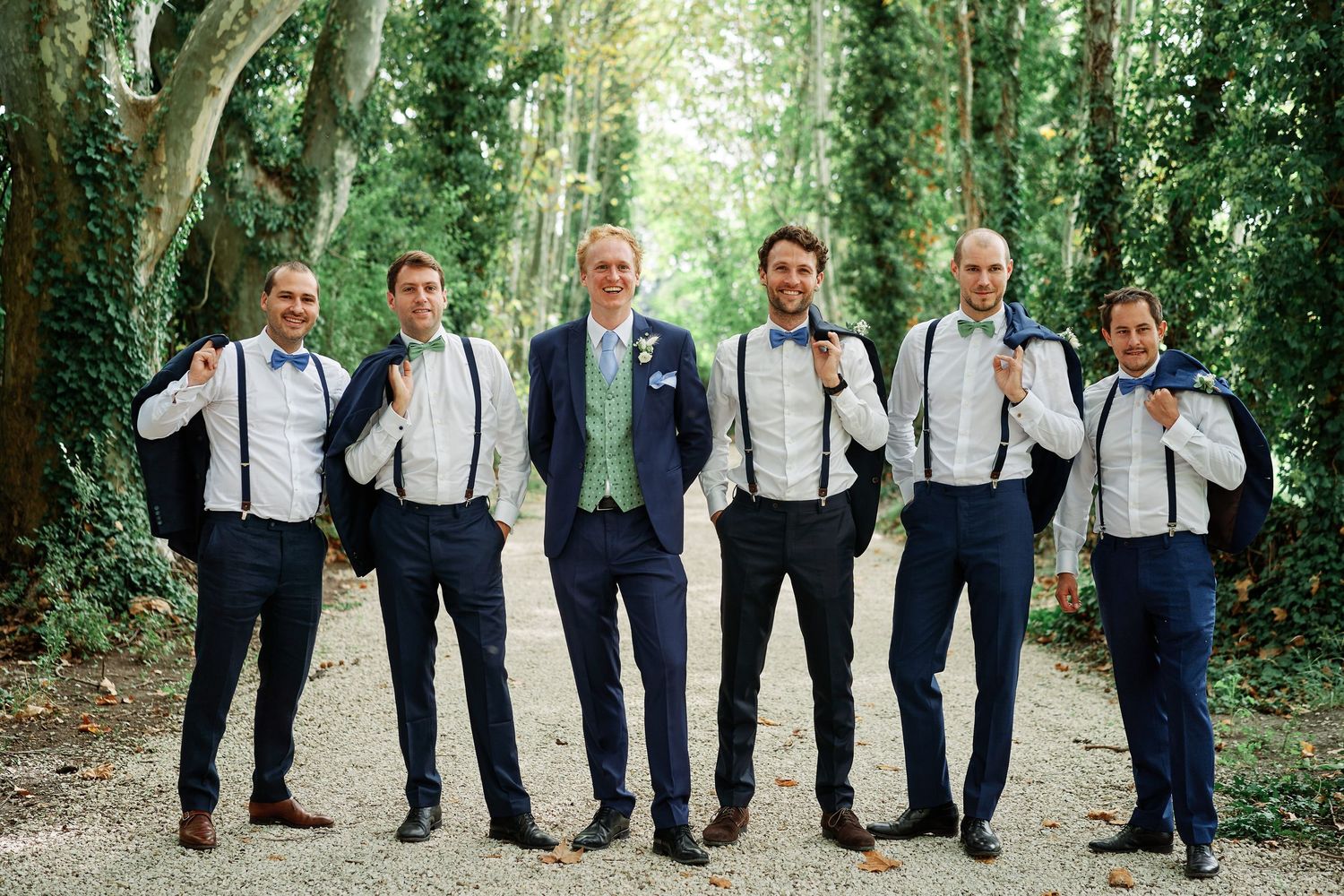 A row of groomsmen in navy suits and suspenders pose together in a wooded outdoor setting for wedding photos.